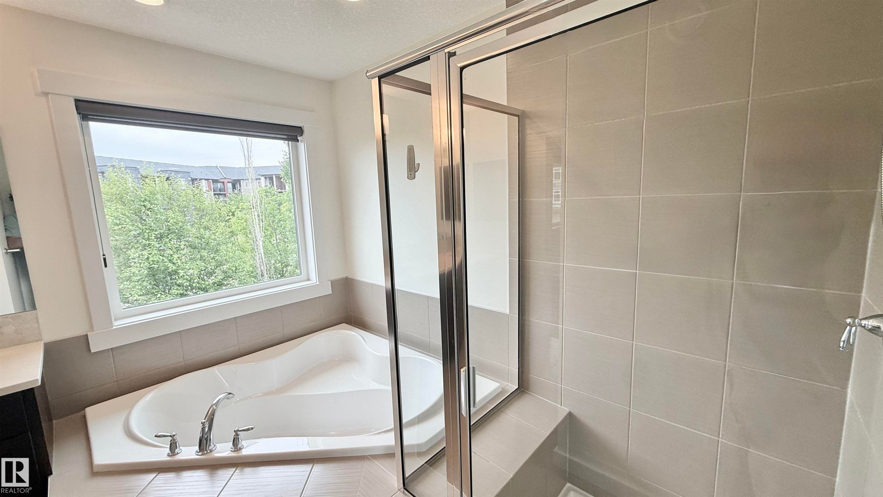 Built-in soaking tub with deck-mounted faucet, glass-enclosed shower, large window with roller shade, and light-toned wall tiling - 3962 Claxton Loop, Edmonton, AB - Indoor Photo Showing Bathroom