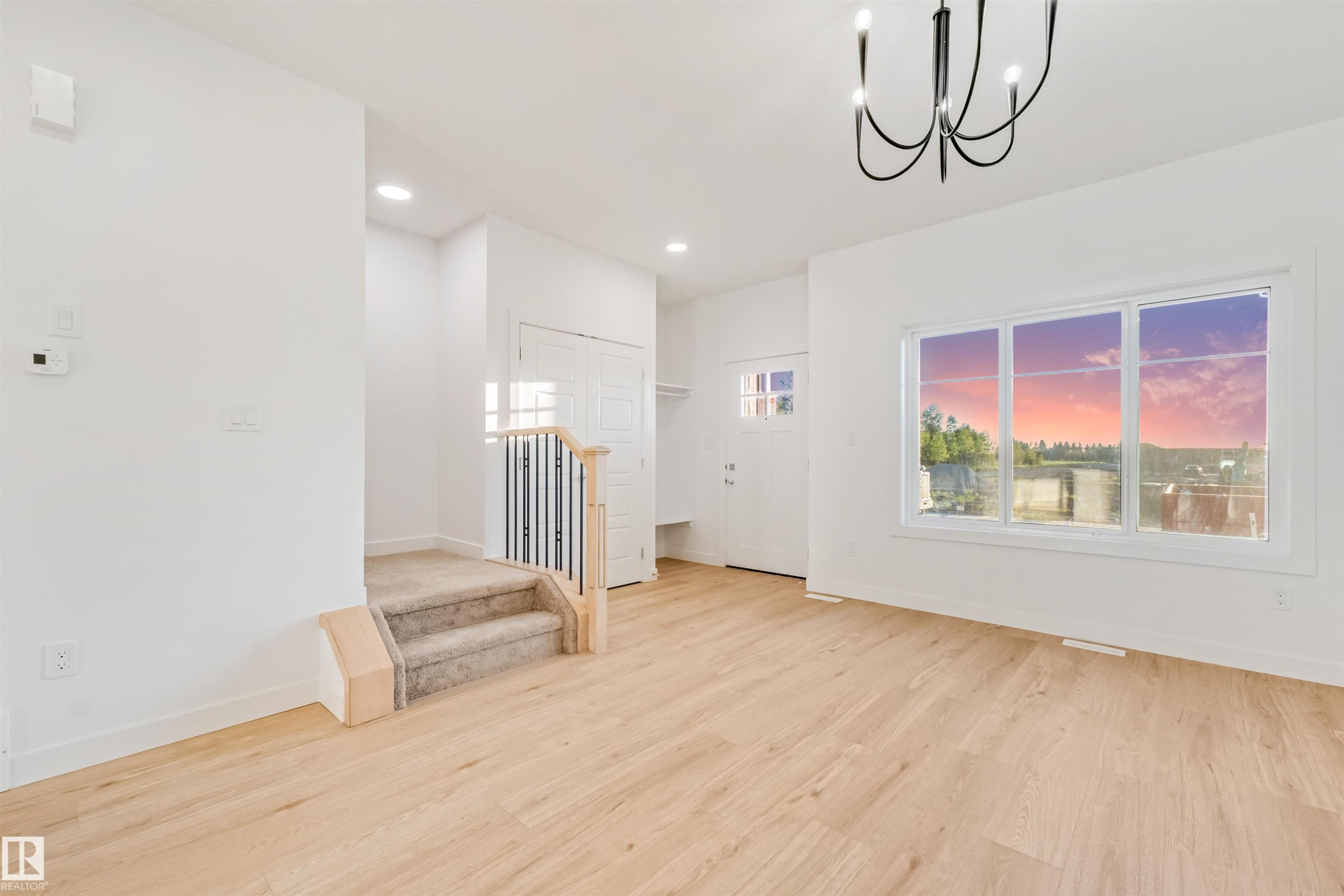 Light wood-finish flooring, white walls, modern chandelier, large multi-pane window, and carpeted stairs with a metal and wood railing - 1511 211 Street Nw, Edmonton, AB