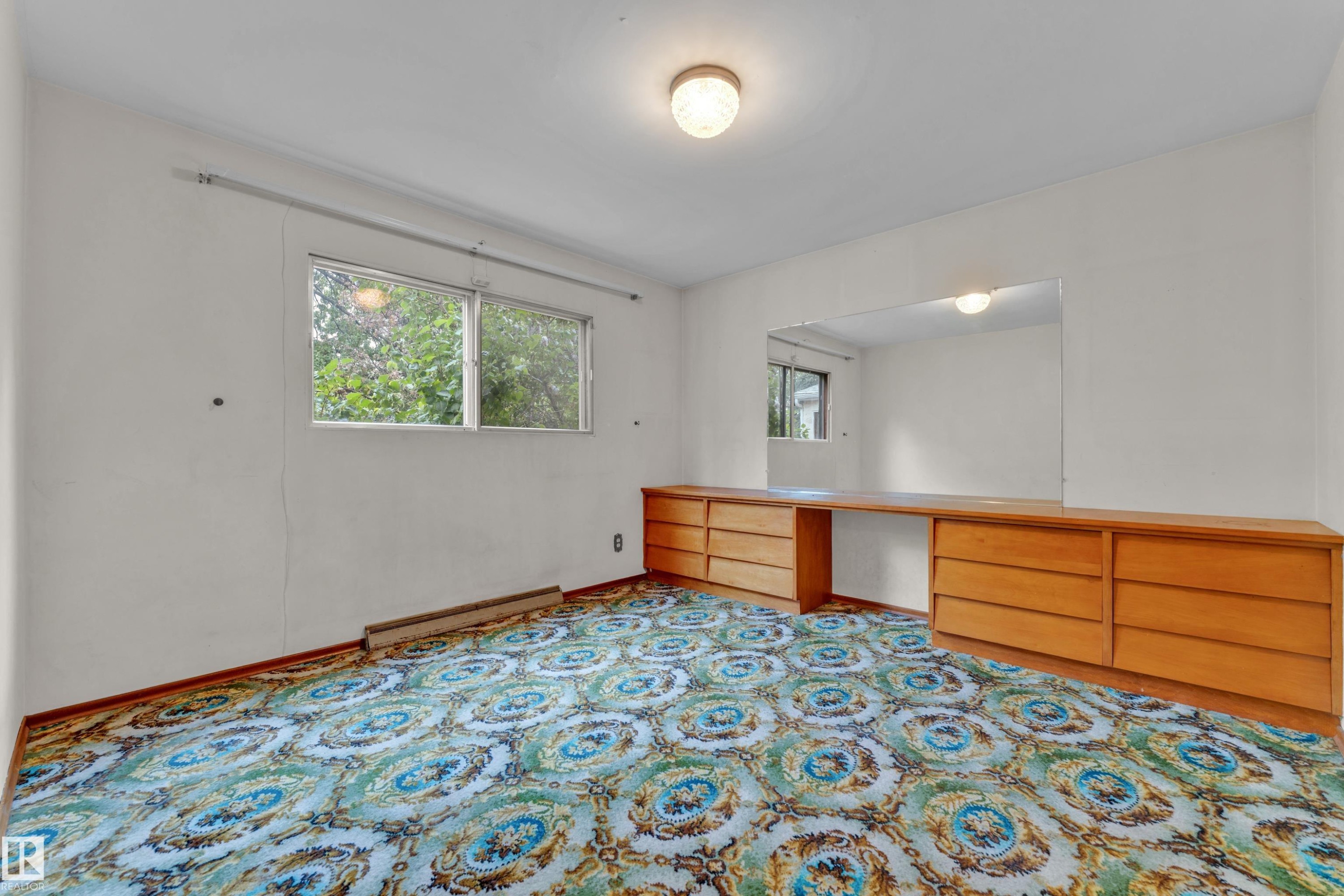 Room featuring a window with exterior foliage, patterned carpet flooring, and a built-in wood-finish vanity with drawer storage - 10819 54 Avenue, Edmonton, AB