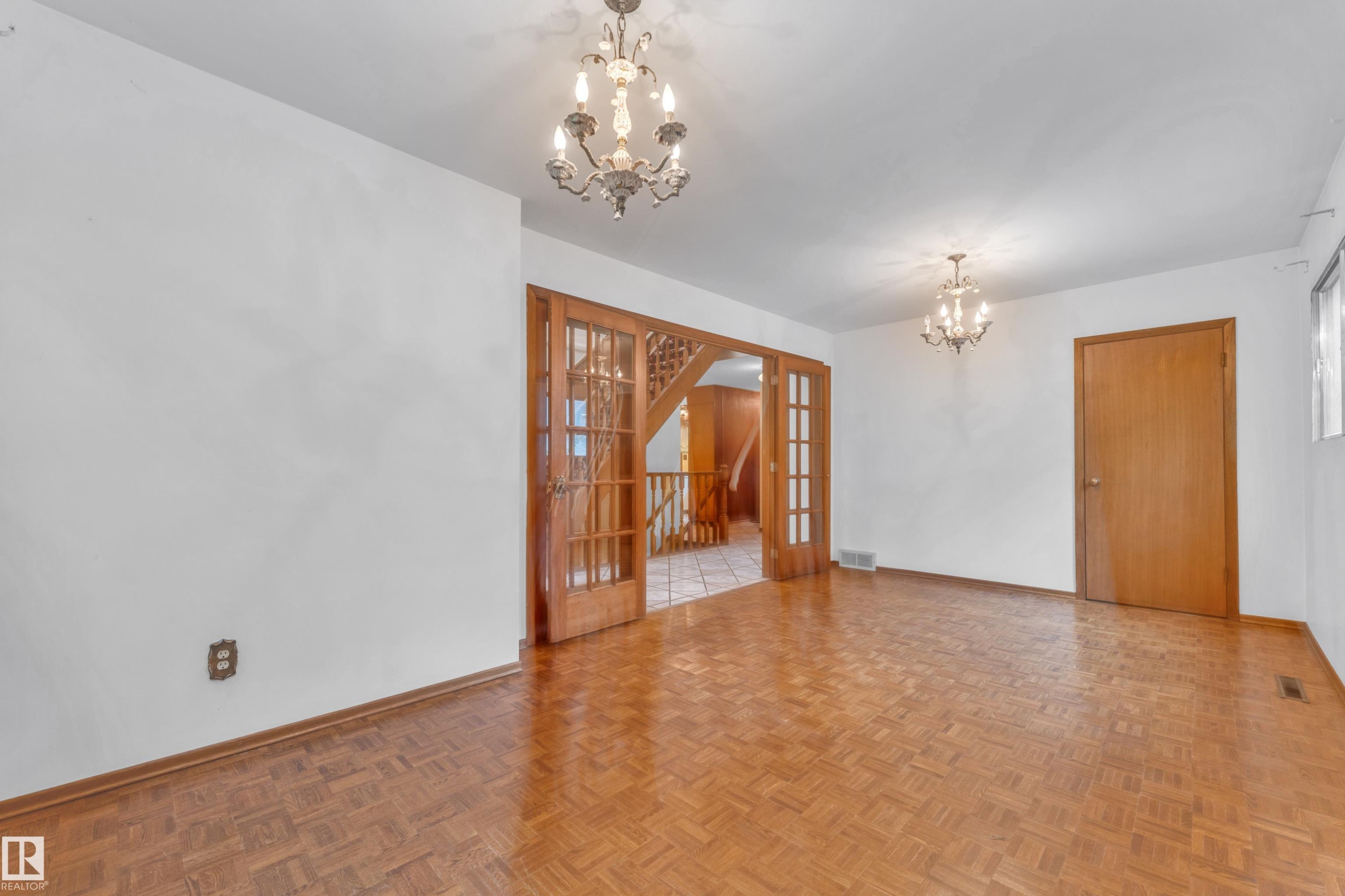 Spacious room with parquet wood-finish flooring, white walls, and two ornate chandeliers - 10819 54 Avenue, Edmonton, AB