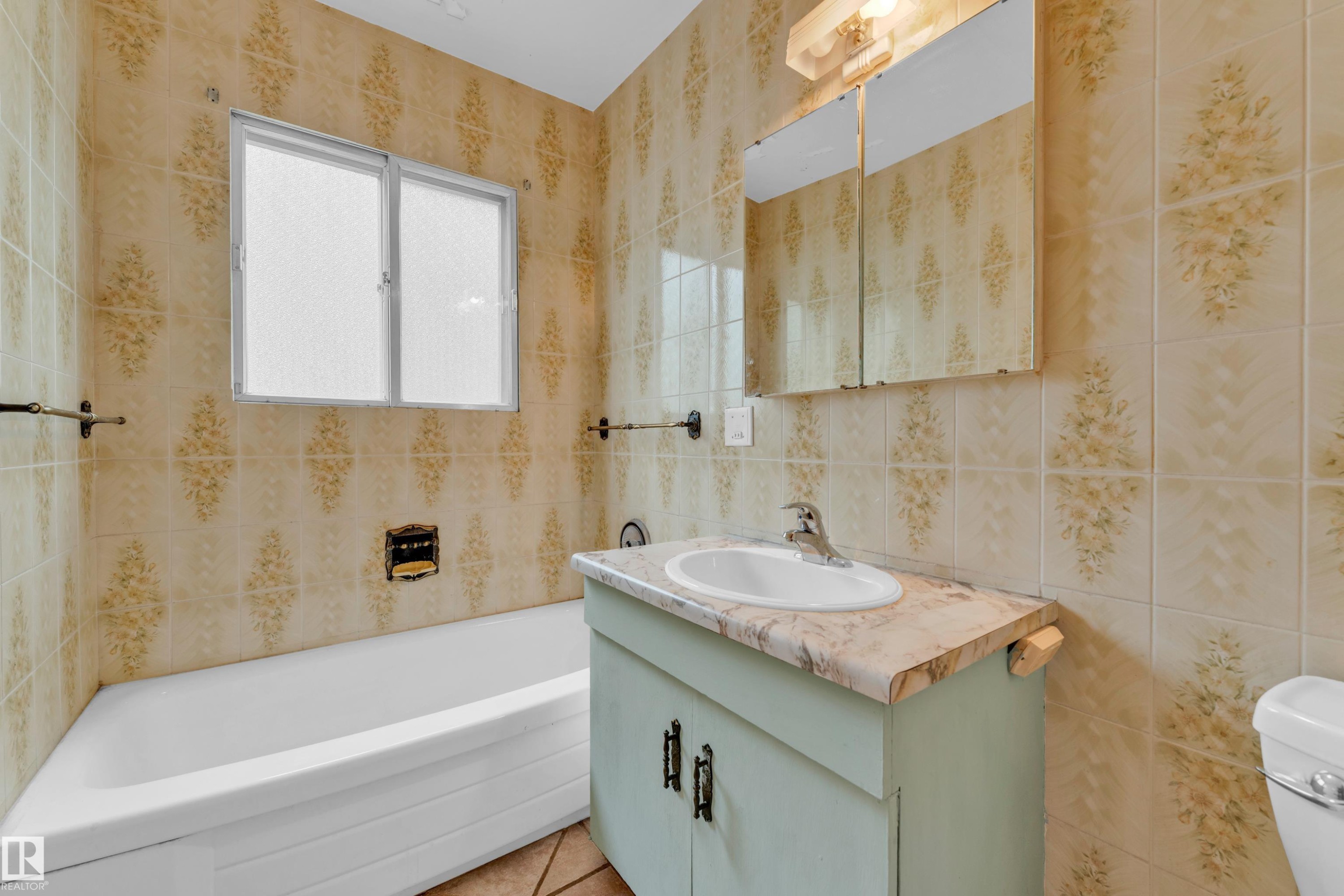 Bathroom featuring patterned wall tiles, a white bathtub, a window with obscure glass, a vanity with a counter-mounted sink, and a mirrored medicine cabinet - 10819 54 Avenue, Edmonton, AB