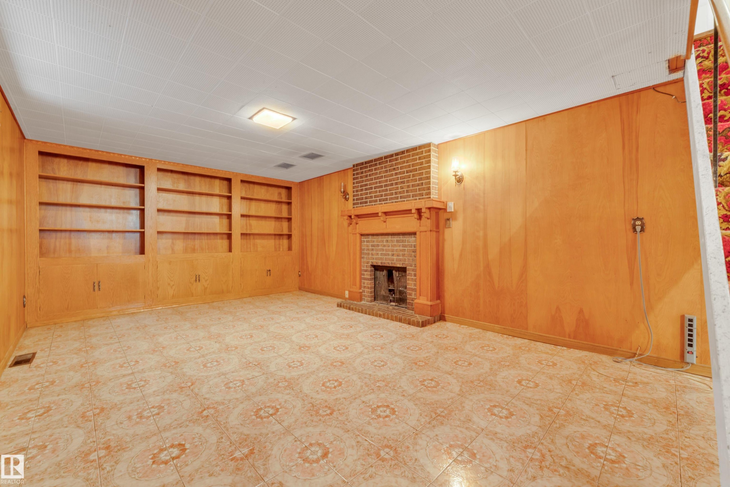 Spacious room featuring wood-paneled walls, built-in shelving with lower cabinetry, and a brick fireplace with a wood mantel - 10819 54 Avenue, Edmonton, AB