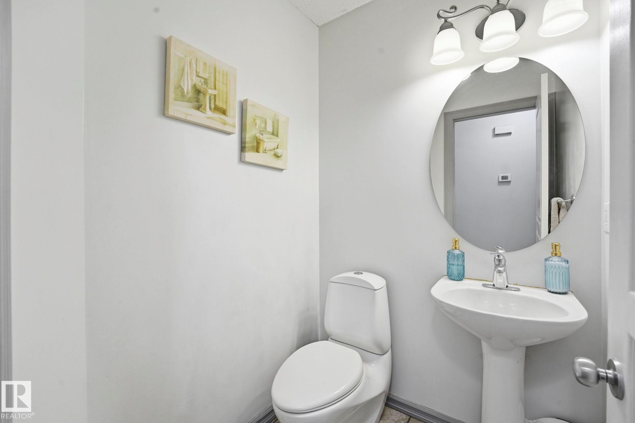 Pedestal sink with chrome faucet, circular mirror, and three-light vanity fixture - 3231 28A Avenue Nw, Edmonton, AB - Indoor Photo Showing Bathroom