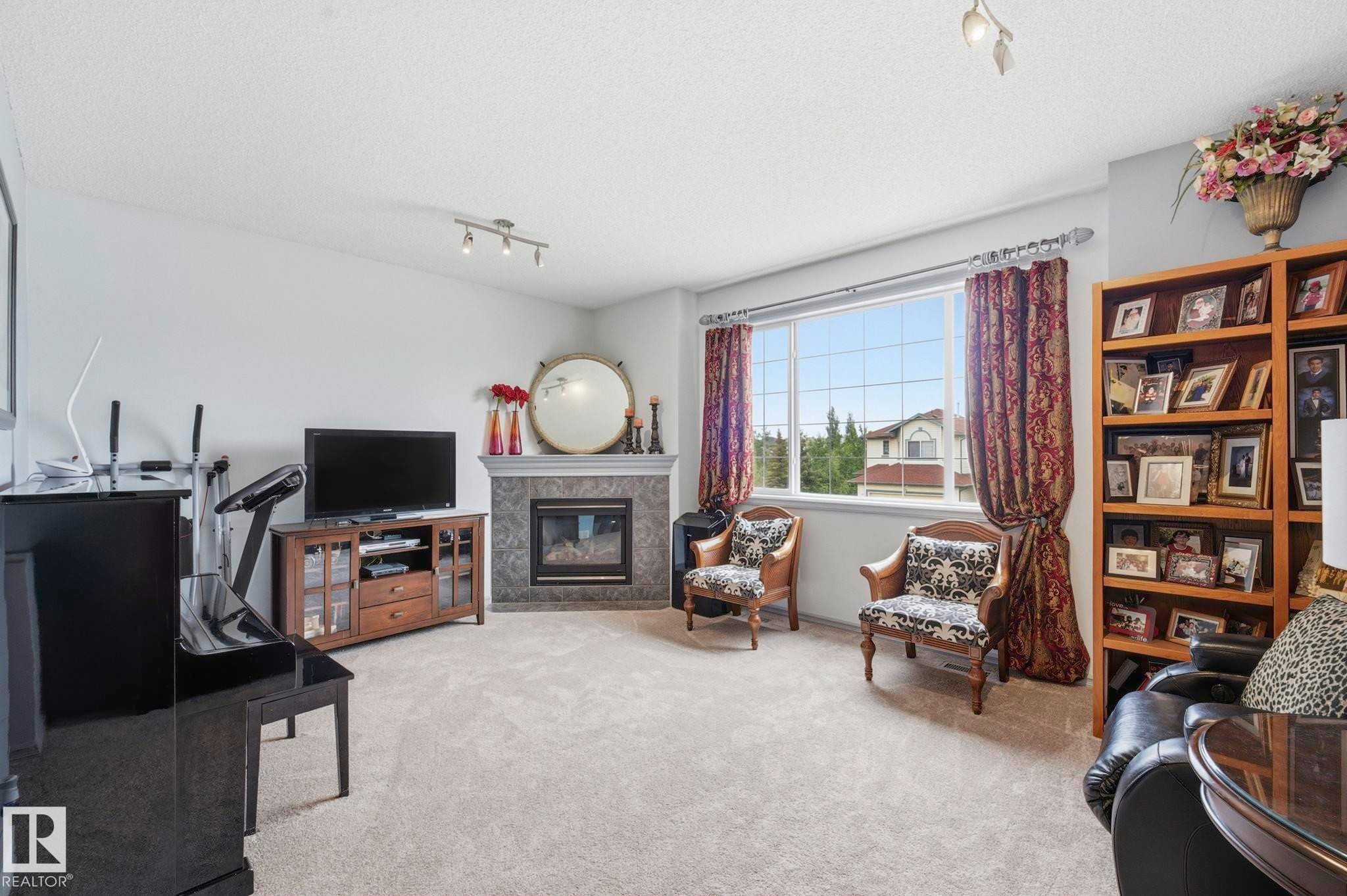 Carpeted living area featuring a corner fireplace with a stone surround and mantel, a large multi-pane window, and a built-in wooden bookshelf - 3231 28A Avenue Nw, Edmonton, AB - Indoor With Fireplace
