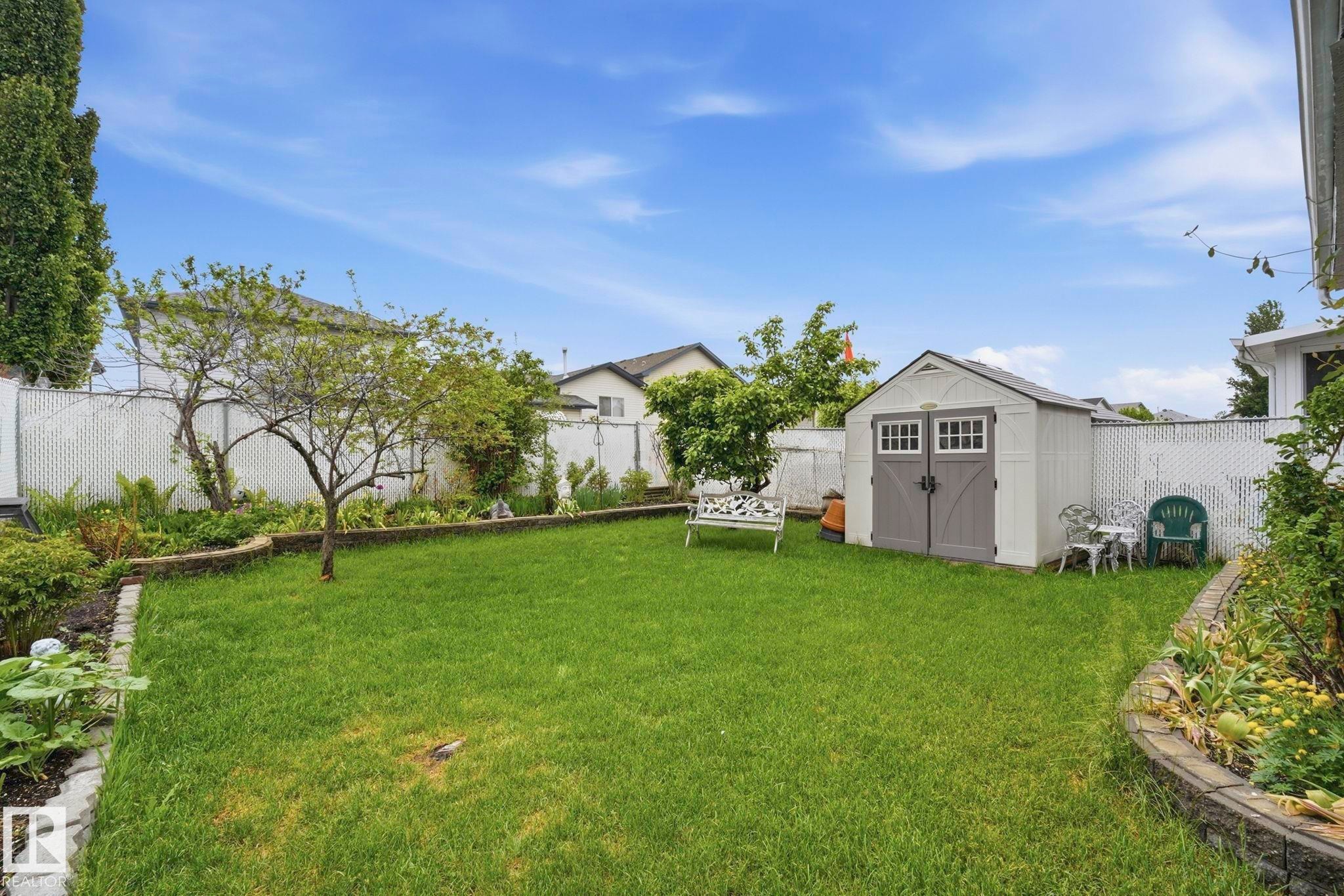 Spacious backyard featuring a manicured lawn, a detached storage shed, raised garden beds with stone edging, mature trees, and a white chain-link fence - 3231 28A Avenue Nw, Edmonton, AB - Outdoor