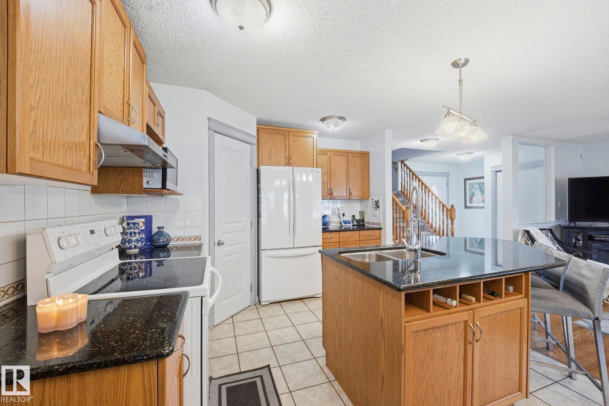 Kitchen featuring light wood cabinetry, black granite countertops, white tile flooring, a functional kitchen island with integrated sink, and an electric range with overhead microwave - 3231 28A Avenue Nw, Edmonton, AB - Indoor Photo Showing Kitchen With Double Sink