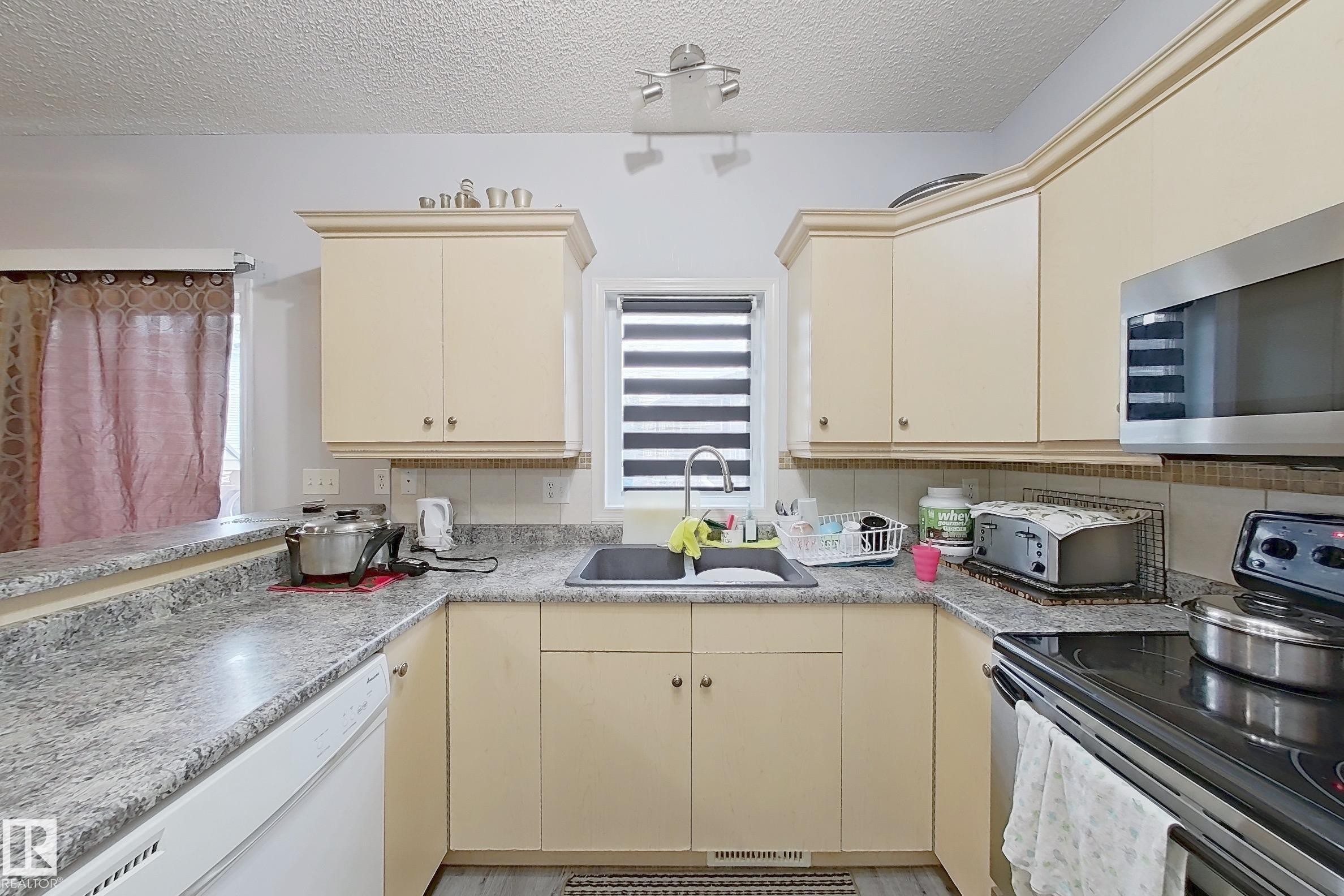 Kitchen featuring cream cabinetry, stainless steel microwave, integrated dishwasher, dark composite sink, and grey flecked countertops - 41 5102 30 Avenue, Beaumont, AB