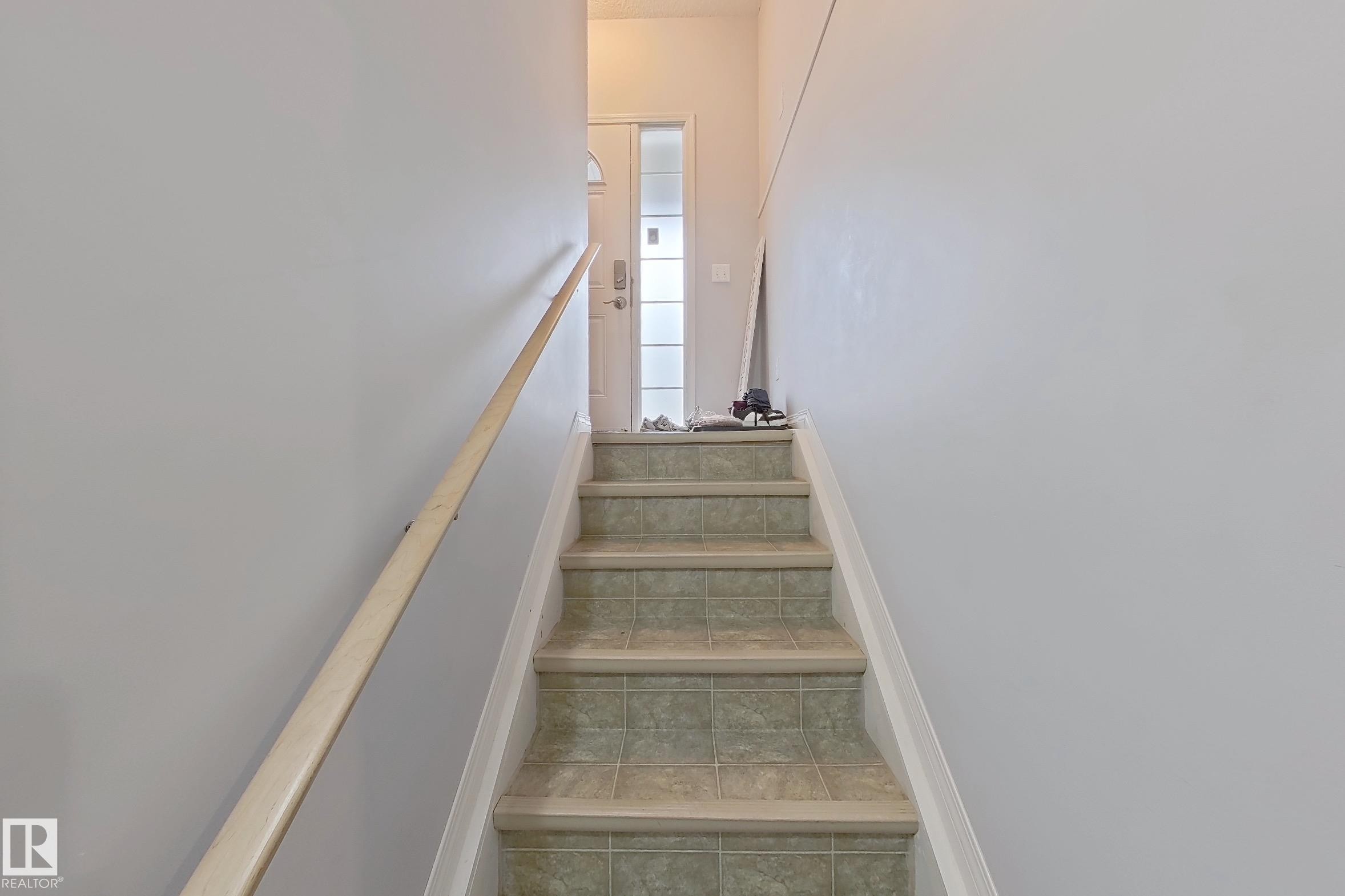 Staircase featuring tiled treads, light-toned risers, a natural wood handrail, and a frosted glass-paneled entry door - 41 5102 30 Avenue, Beaumont, AB