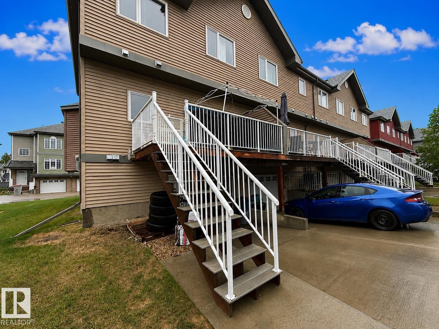 Rear exterior featuring tan siding, a ground-level garage, an upper-level deck with white railings, and an exterior staircase with white railings - 41 5102 30 Avenue, Beaumont, AB