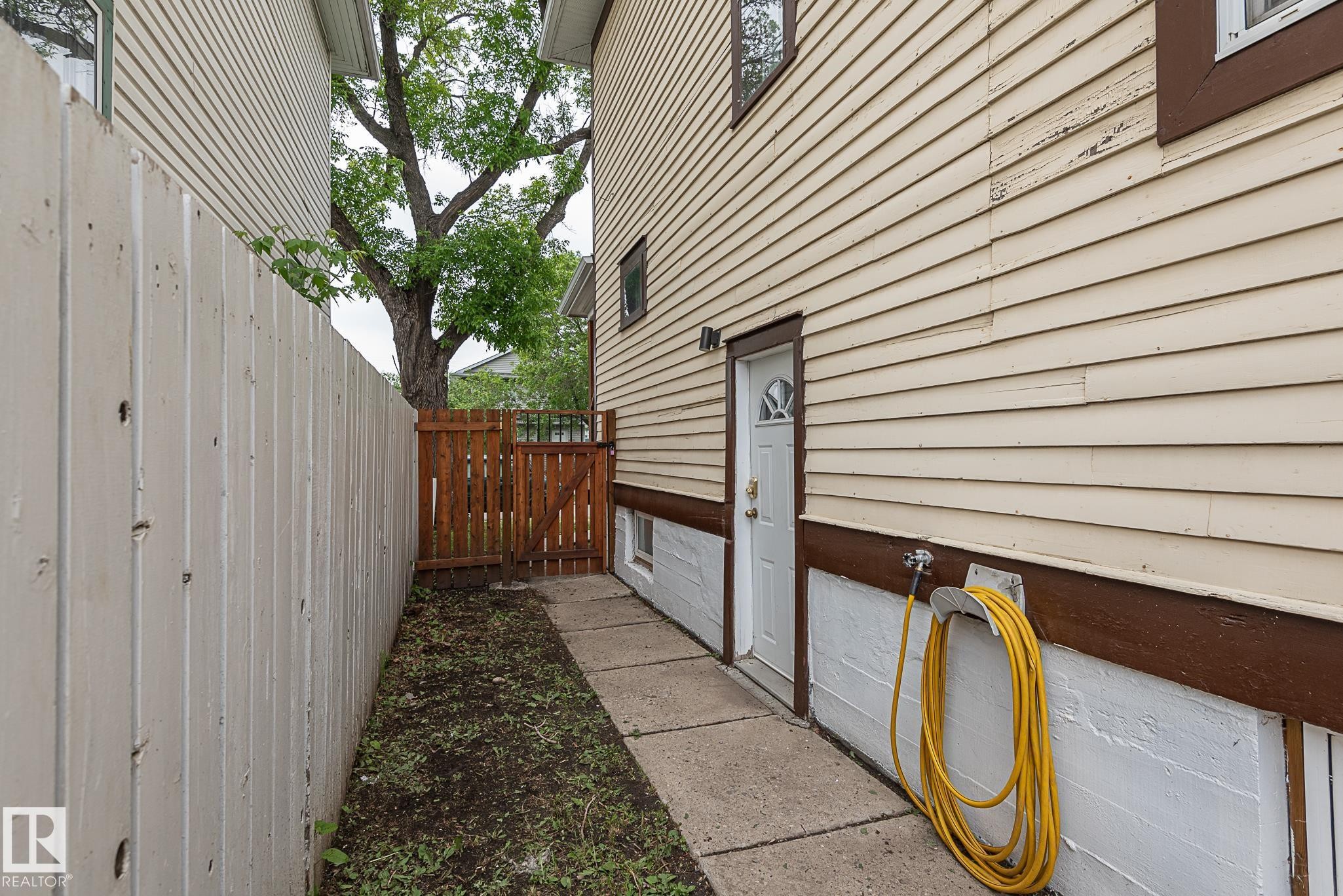 Side exterior featuring horizontal siding, a white entry door with a decorative glass insert, a concrete pathway, a wooden privacy fence, and a wooden gate - 9623 110A Avenue, Edmonton, AB