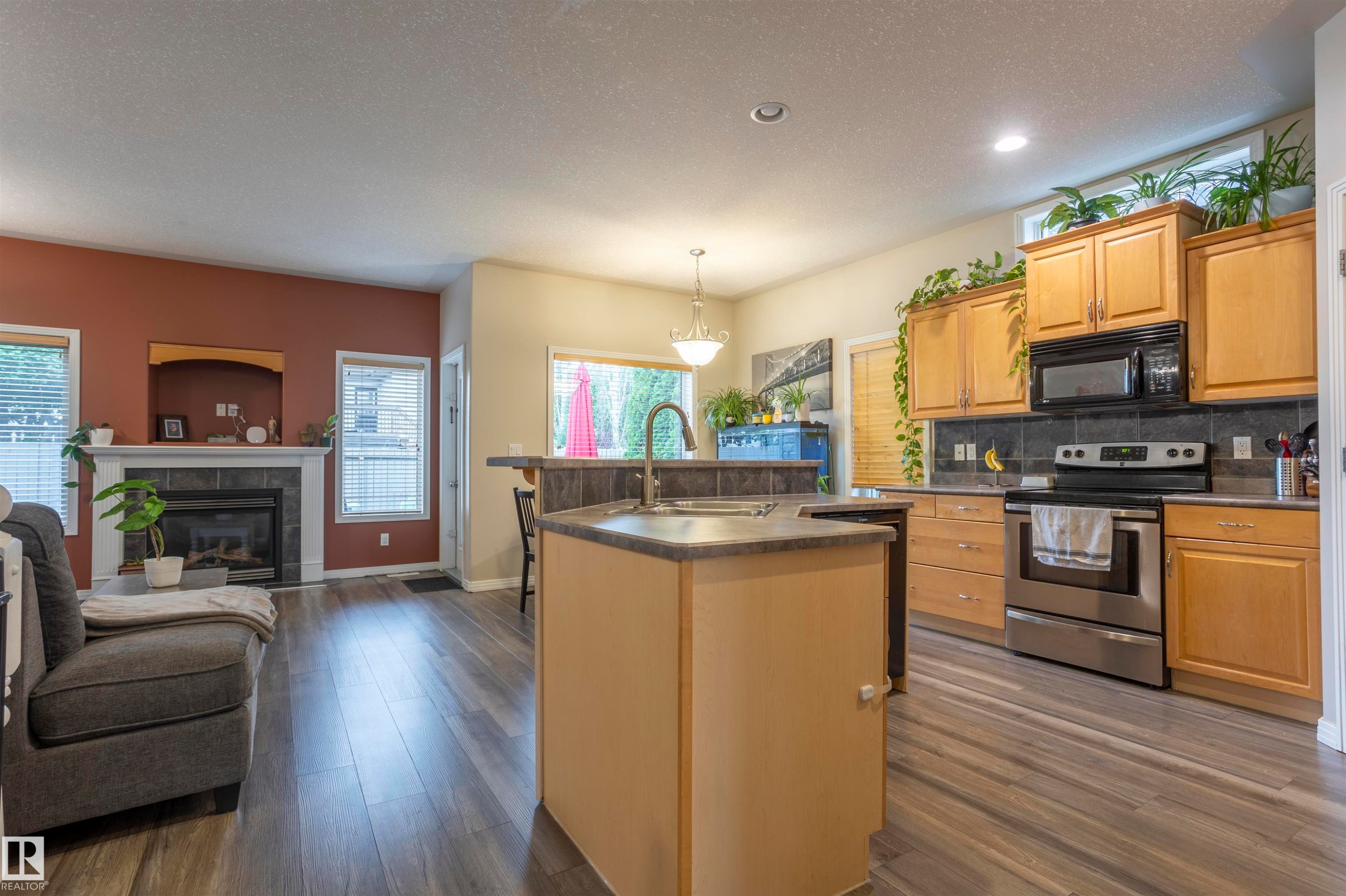 Open-concept living area featuring wood-finish flooring, a kitchen island with sink, stainless steel appliances, a fireplace with a mantel, and multiple windows - 1441 Highwood Boulevard, Devon, AB