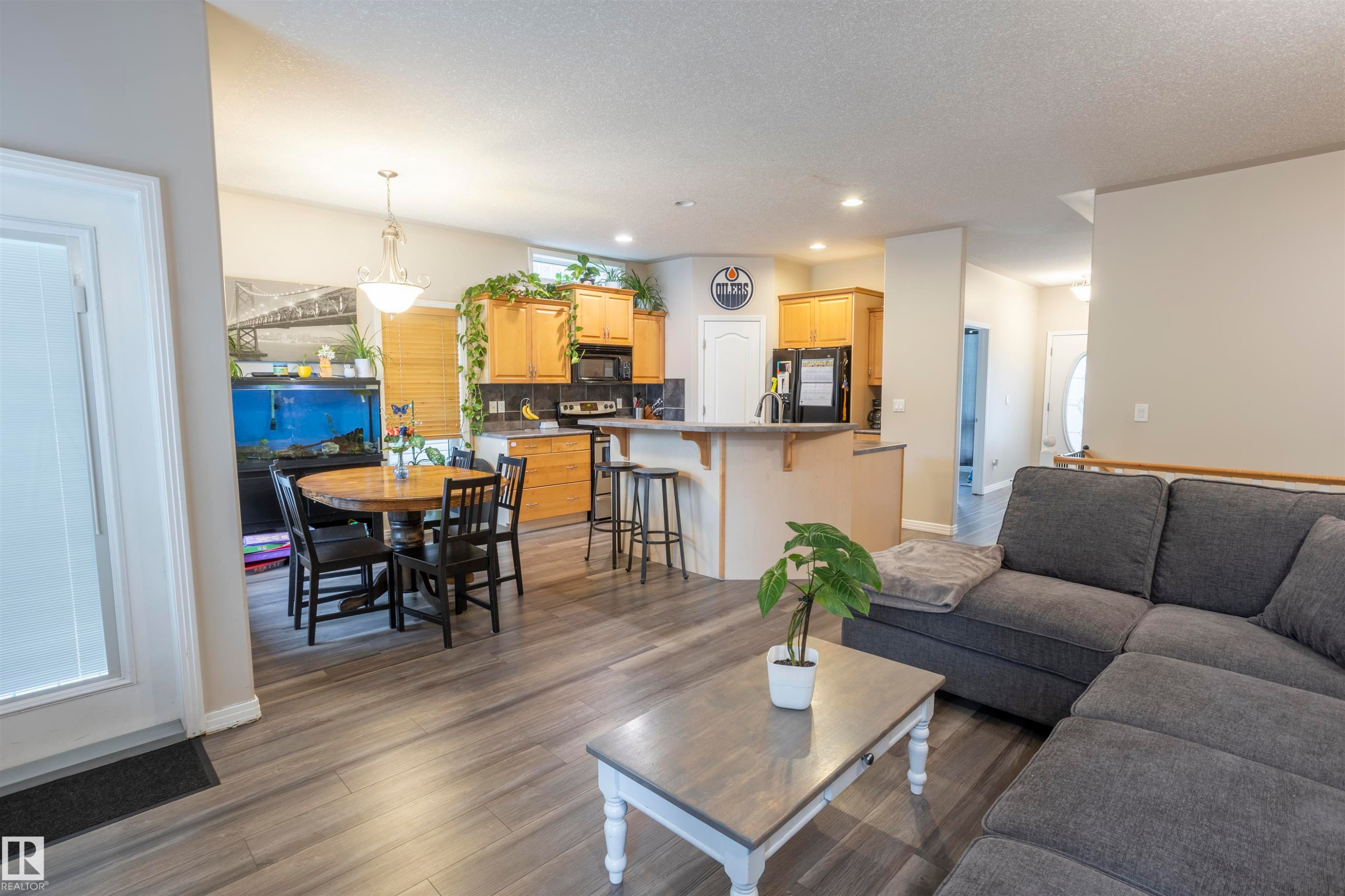 Open-concept living space with wood-finish flooring, a kitchen featuring light wood cabinetry and a breakfast bar, and a dining area with an overhead chandelier - 1441 Highwood Boulevard, Devon, AB