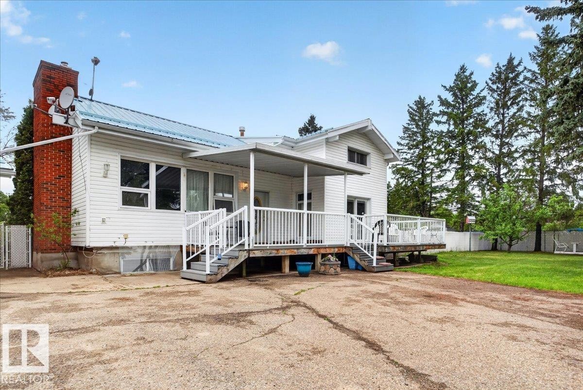 White siding exterior with a blue metal roof, featuring a red brick chimney, an elevated wood deck with white railings, and a covered porch area - 140 50047 Rge Rd 244, Rural Leduc County, AB