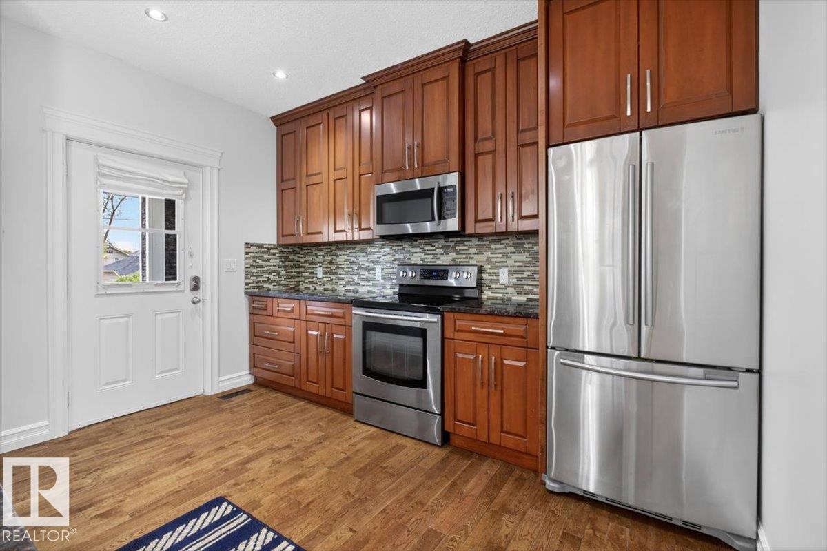 Kitchen featuring wood-finish flooring, stainless steel appliances, rich wood cabinetry, a mosaic tile backsplash, and recessed lighting - 10958 88 Avenue, Edmonton, AB