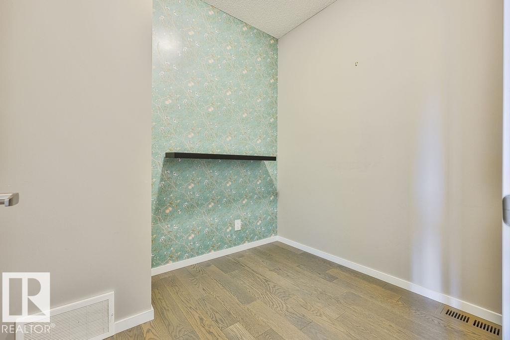Room featuring a decorative accent wall with light green patterned wallpaper, wood-finish flooring, white baseboards, and a black floating shelf - 2031 207 Street, Edmonton, AB