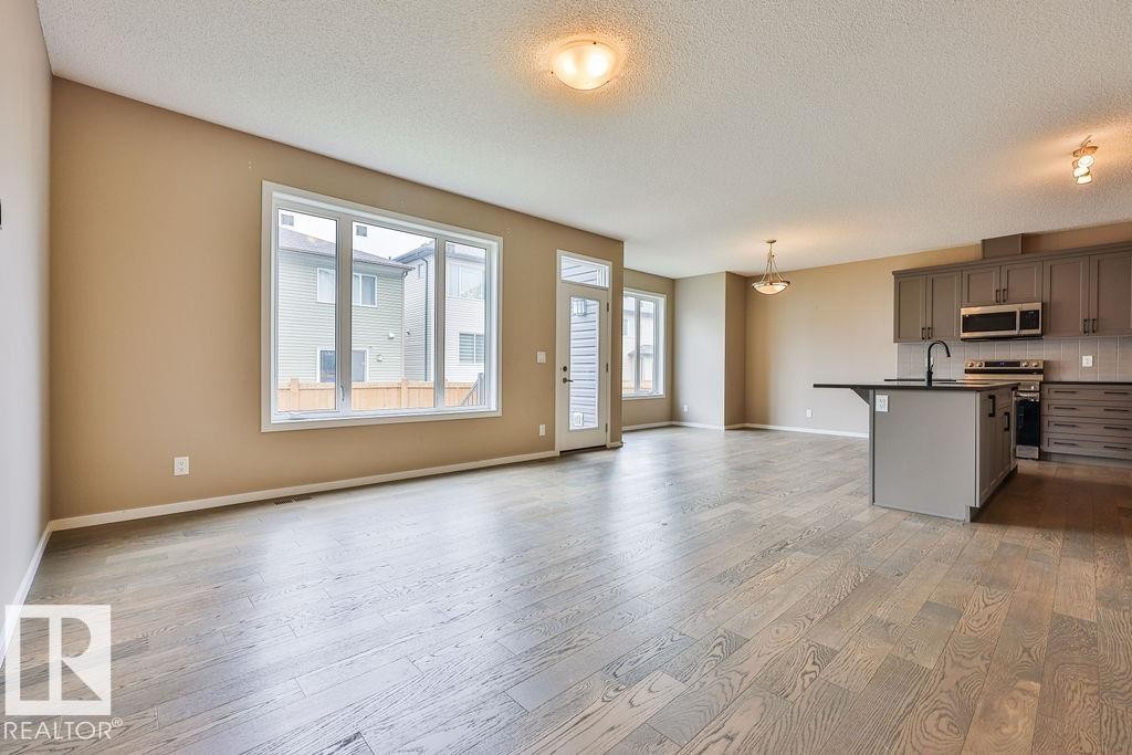 Open-concept living space featuring wood-finish flooring, large windows, a kitchen island with seating, dark shaker-style cabinetry, and stainless steel appliances - 2031 207 Street, Edmonton, AB