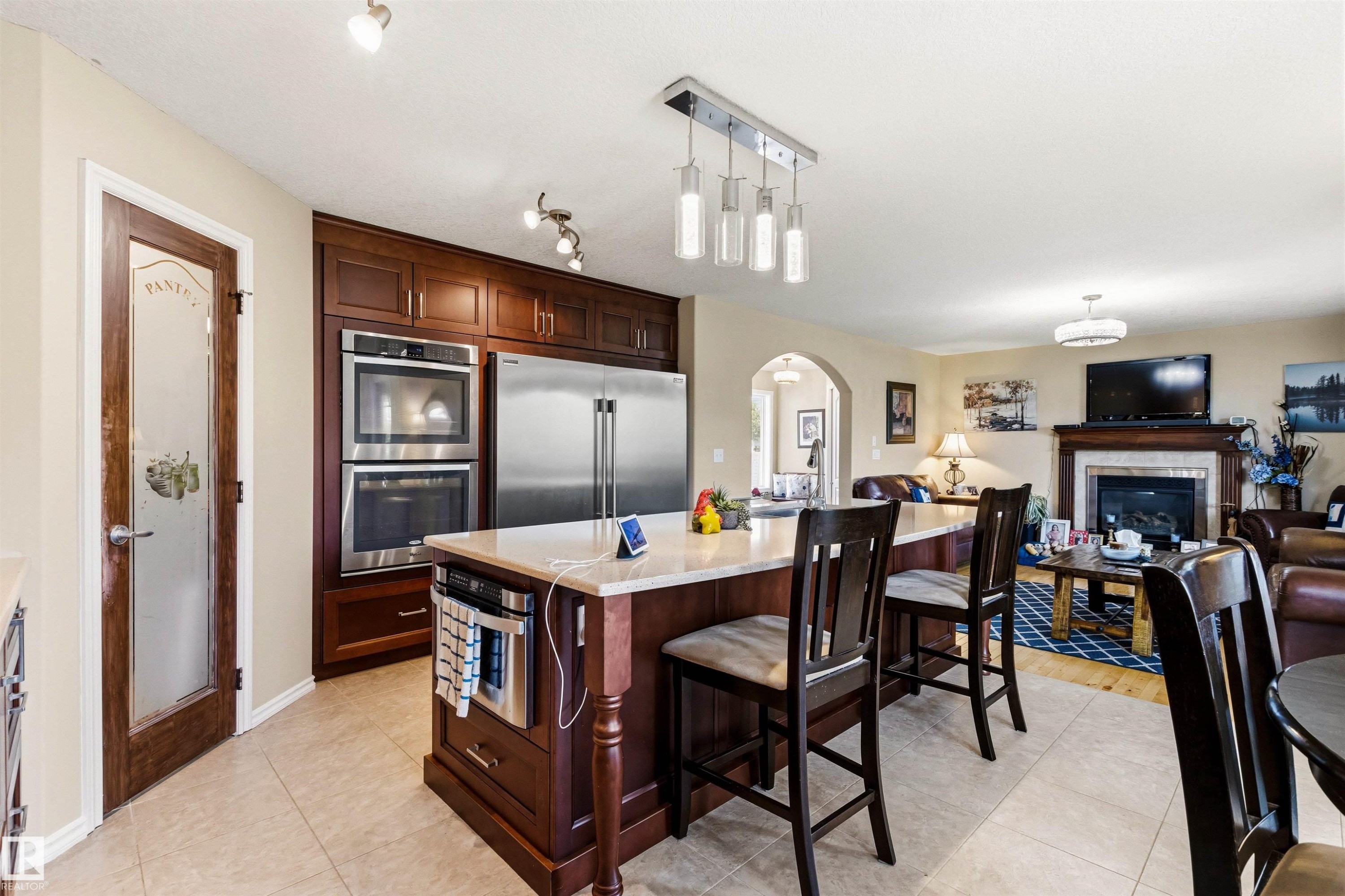 Kitchen featuring a center island with countertop seating, built-in stainless steel appliances, dark wood cabinetry, and a pantry door with frosted glass - 32 Lamplight Bay, Spruce Grove, AB