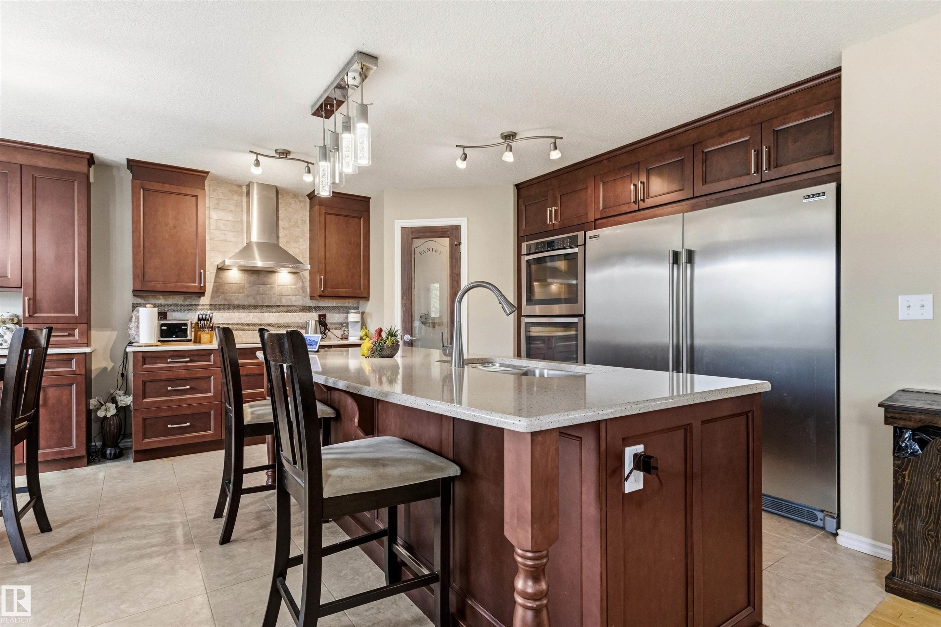 Expansive kitchen featuring a large central island with seating, ample cherry wood cabinetry, stainless steel appliances, a tile backsplash, and overhead pendant lighting - 32 Lamplight Bay, Spruce Grove, AB