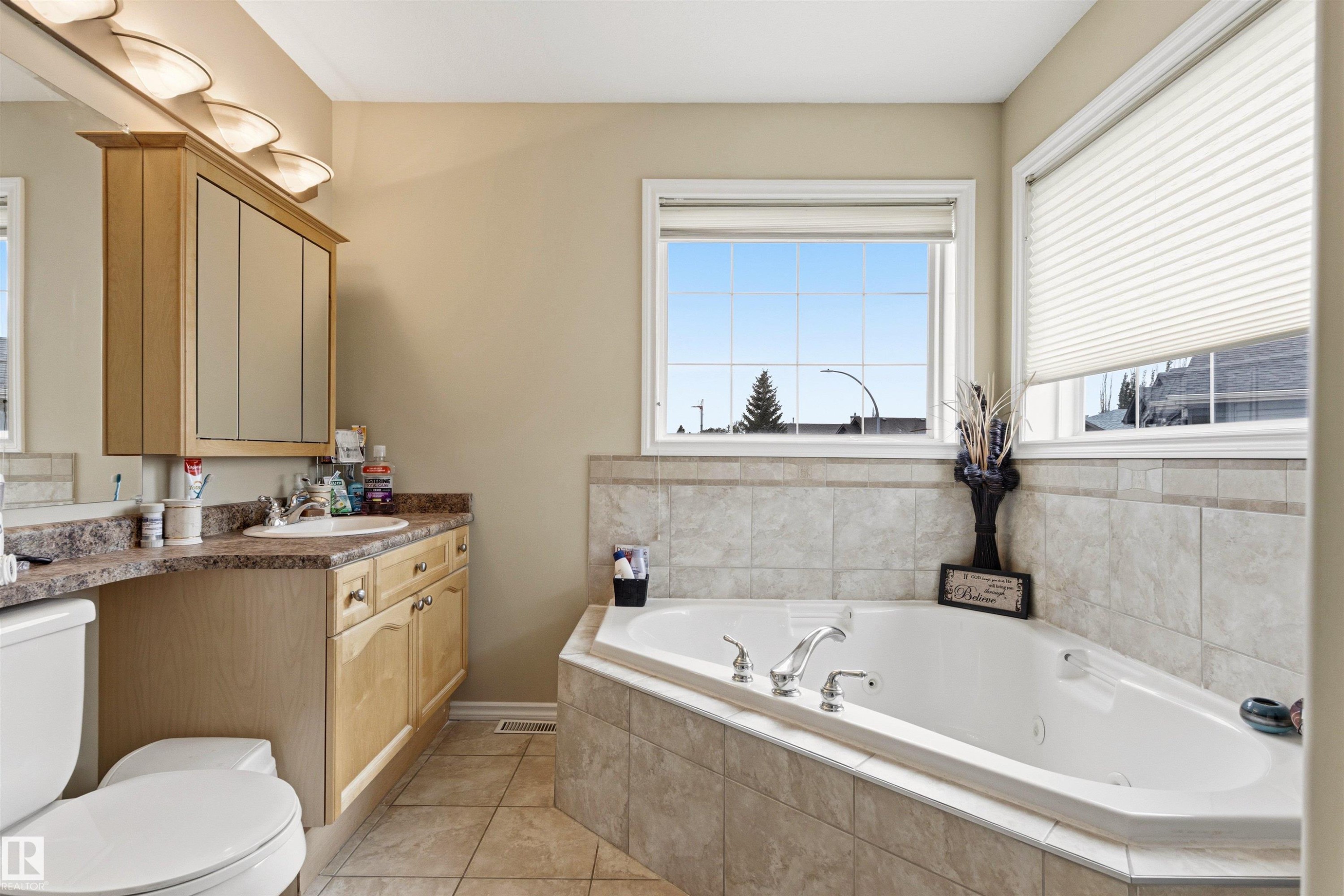 Spacious bathroom featuring a corner soaking tub with tiled surround, a light wood-finish vanity with an integrated sink and ample counter space, multiple windows, and ceramic tile flooring - 32 Lamplight Bay, Spruce Grove, AB