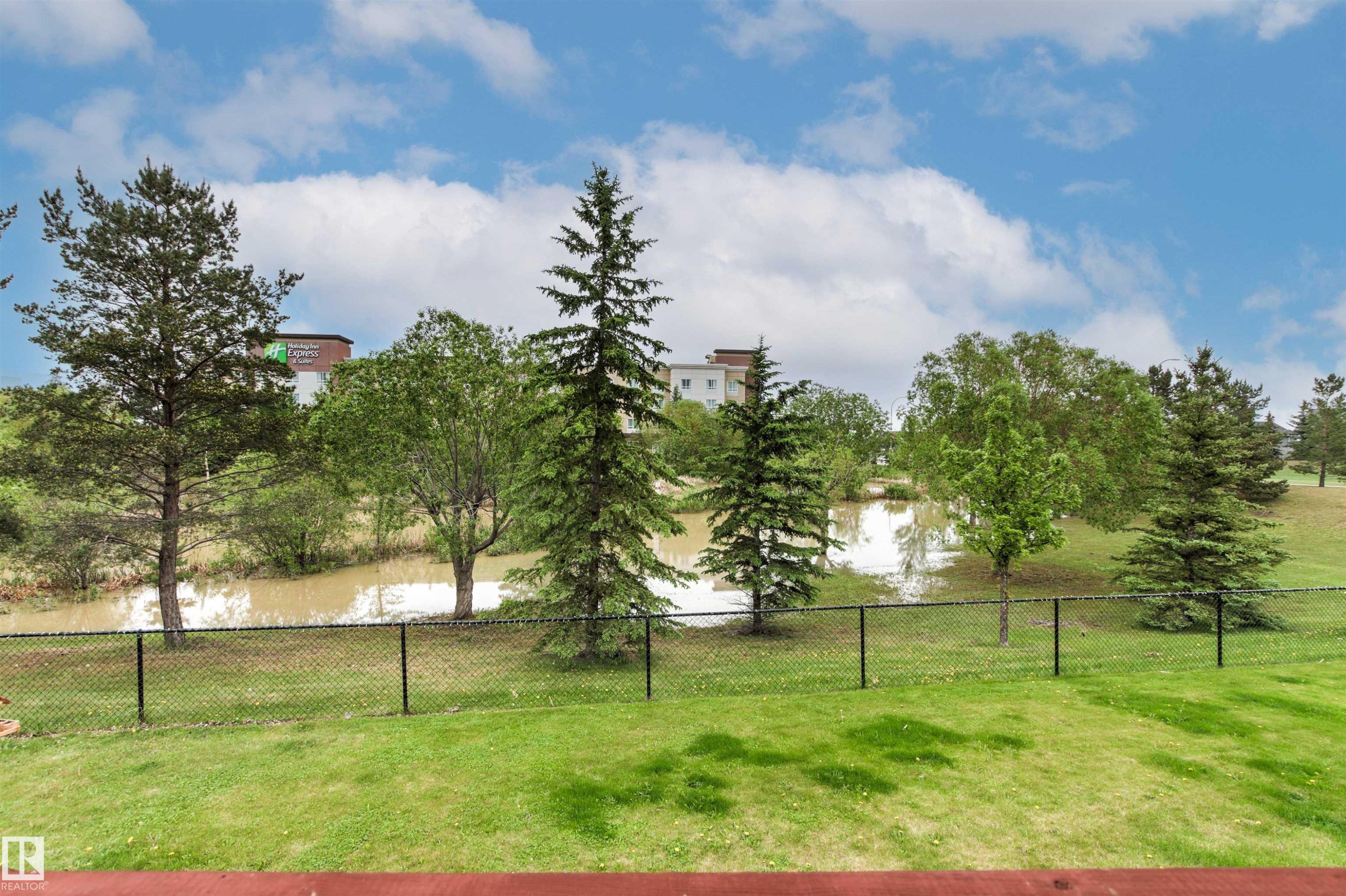 Green lawn area bordered by a black chain-link fence, offering a park-like setting with a body of water and mature trees - 32 Lamplight Bay, Spruce Grove, AB