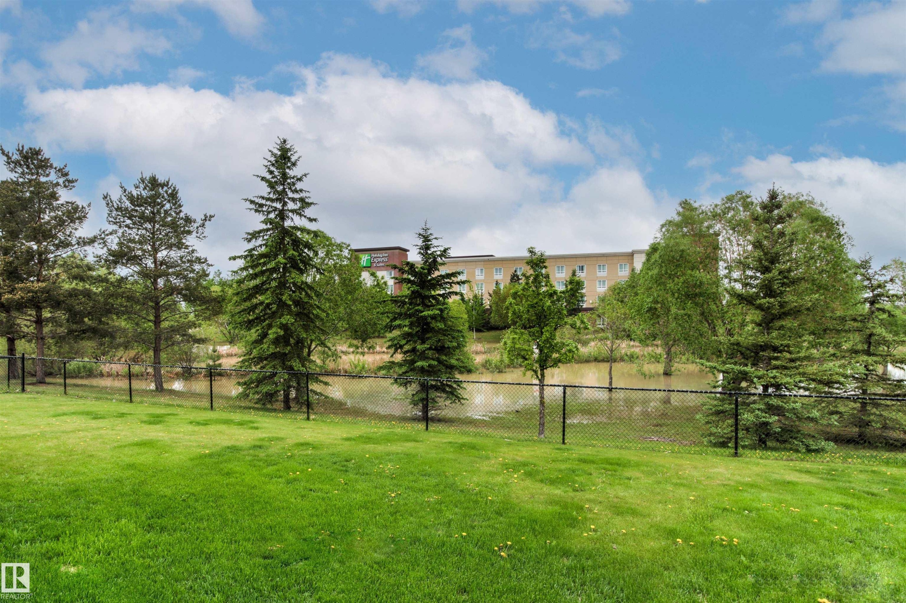Expansive green lawn with a black chain-link fence, mature evergreen trees, and a visible pond - 32 Lamplight Bay, Spruce Grove, AB