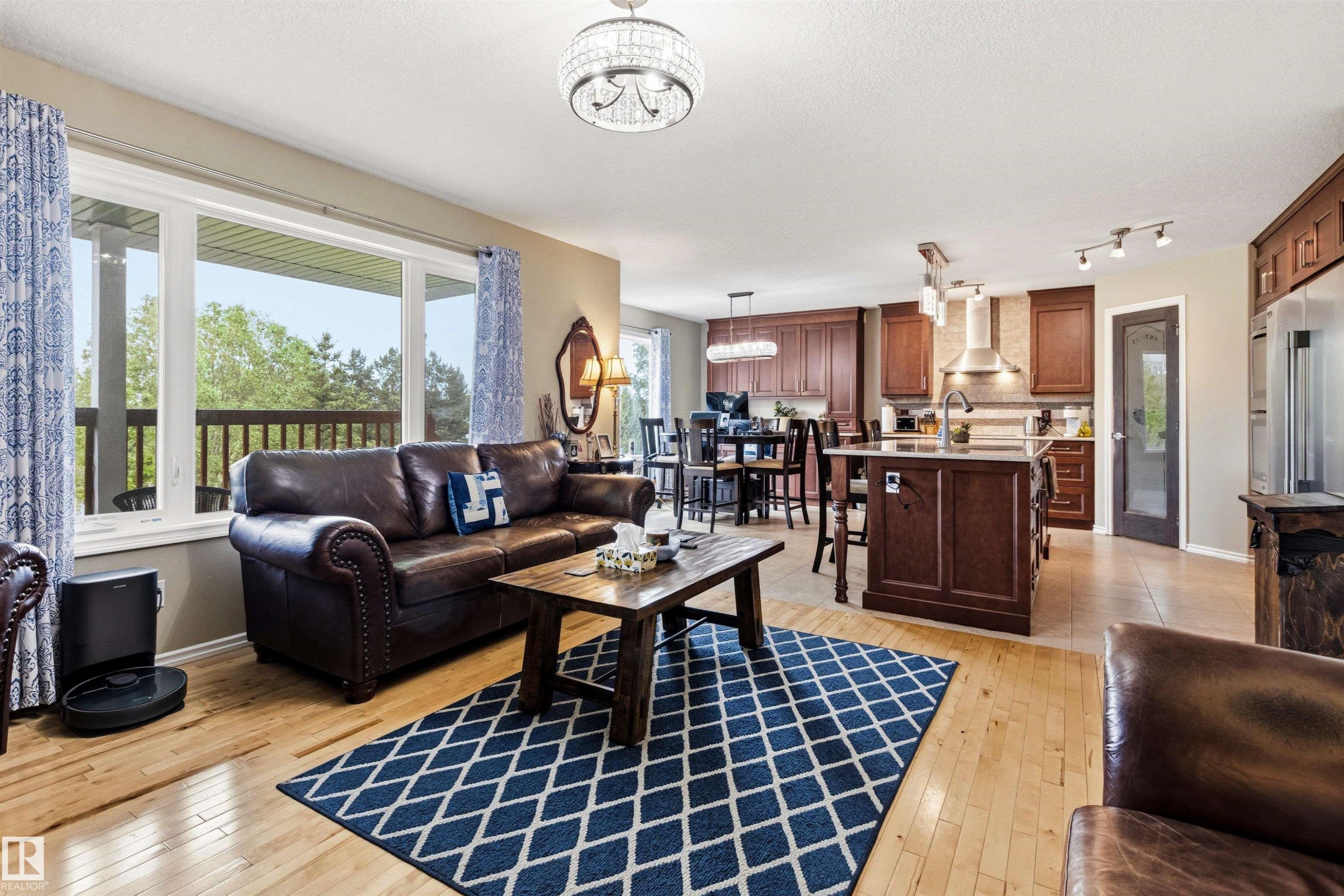Open-concept living area featuring light wood-finish flooring, a large window overlooking a wooden deck and trees, an island kitchen with dark wood cabinetry, stainless steel appliances, and a dining area with a chandelier - 32 Lamplight Bay, Spruce Grove, AB