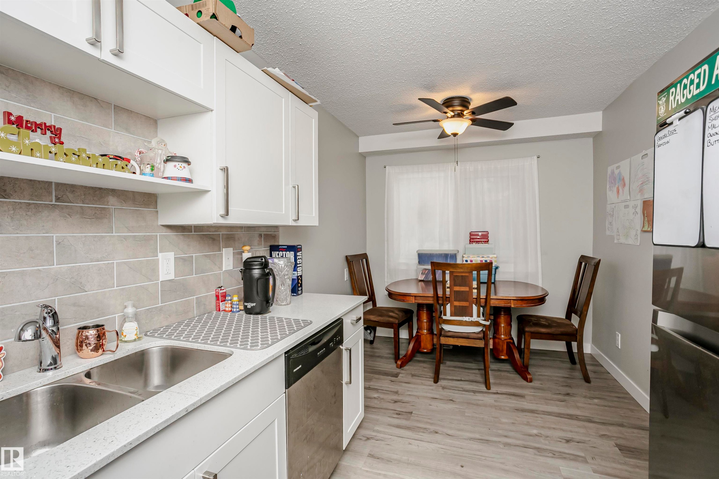 Kitchen and dining area featuring white cabinetry with silver hardware, light grey subway tile backsplash, a double basin stainless steel sink, wood-finish flooring, and a ceiling fan with integrated lighting - 3103 109 Street, Edmonton, AB