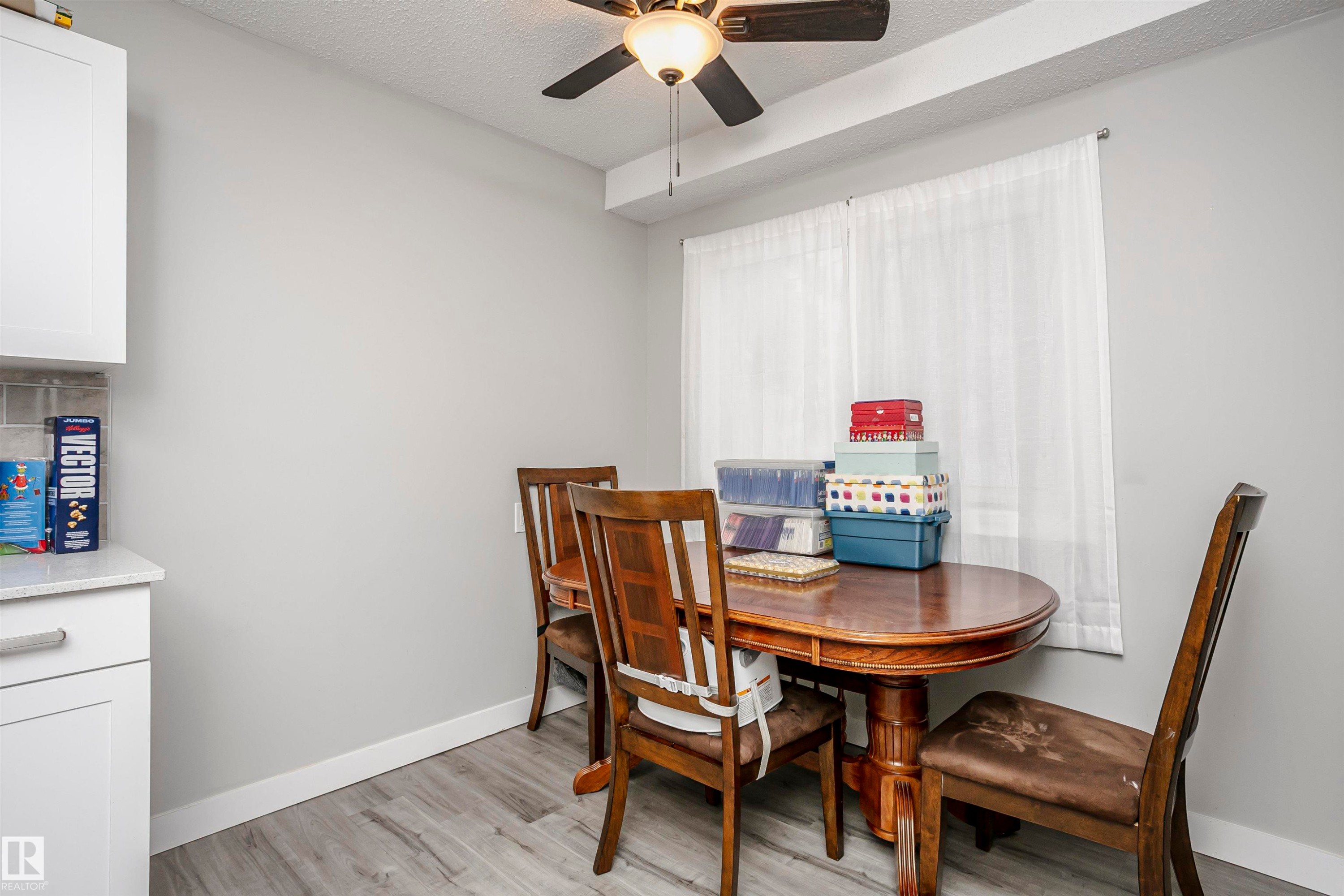 Dining area featuring a contemporary ceiling fan, a large window with sheer white curtains, light gray walls, and wood-finish flooring - 3103 109 Street, Edmonton, AB