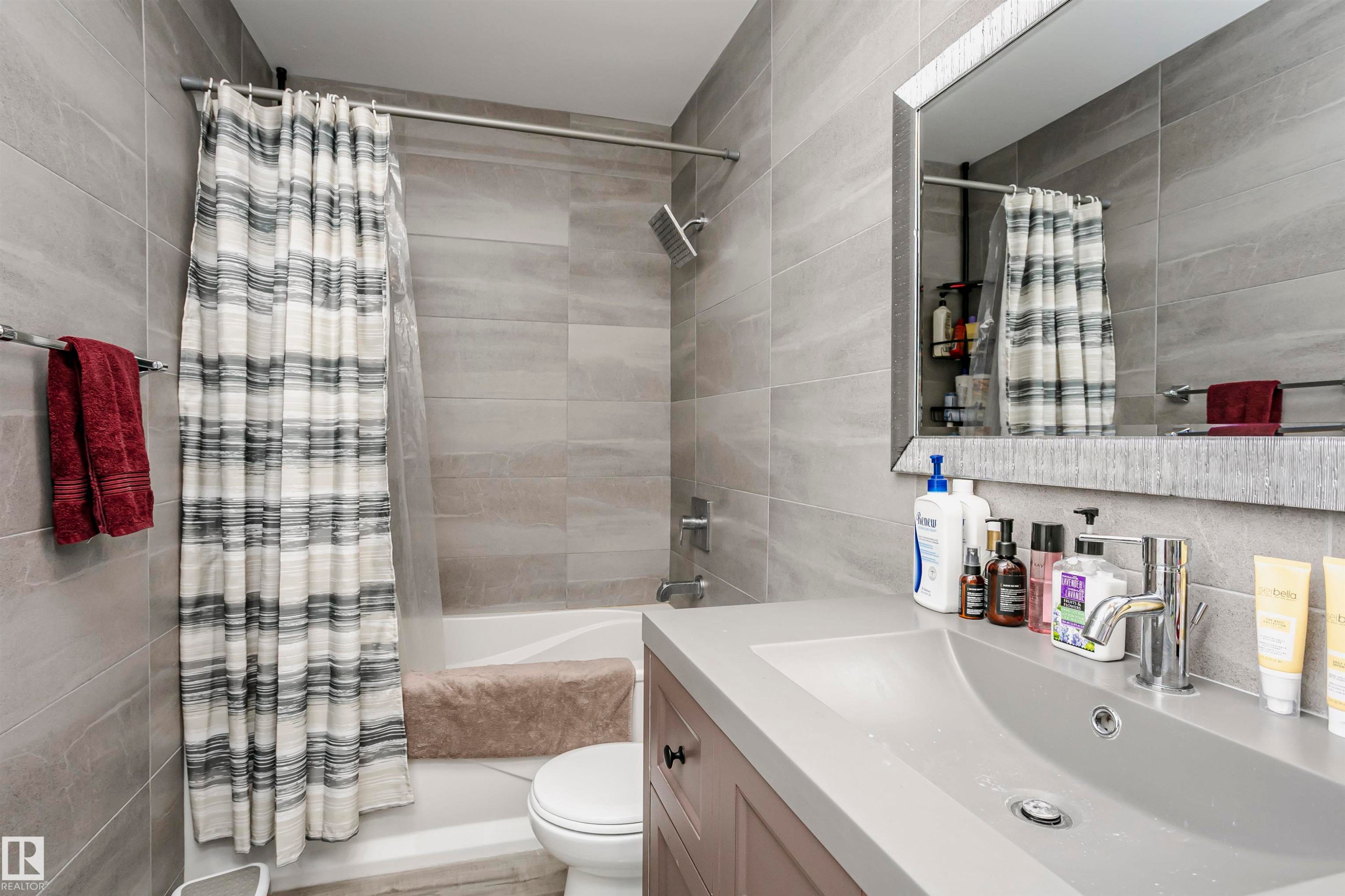 Bathroom featuring a built-in tub with a shower fixture, expansive gray tile surround, and a coordinating vanity with an integrated sink - 3103 109 Street, Edmonton, AB