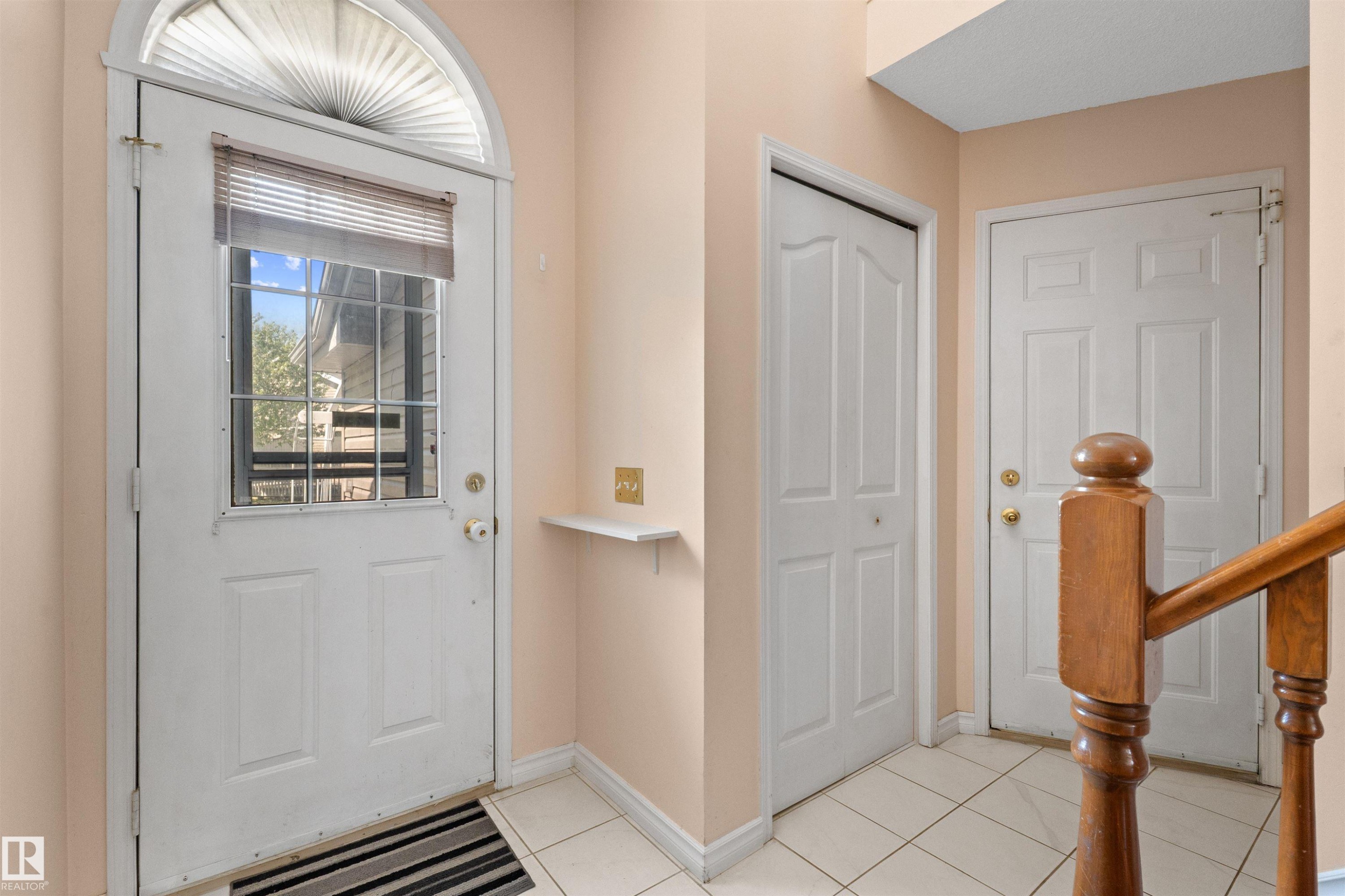 Entryway featuring a white paneled door with glass inserts and an arched transom window, ceramic tile flooring, a built-in shelf, and a light wood staircase banister - 5 20 Deerbourne Drive, St. Albert, AB