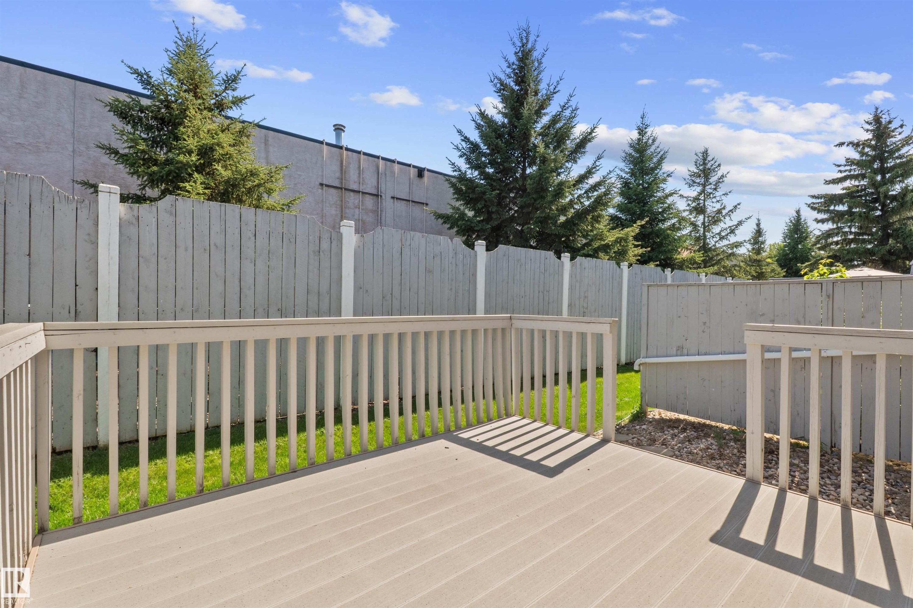 Expansive outdoor deck with composite decking and light-colored railing, featuring a private fenced yard with established evergreen trees and green lawn - 5 20 Deerbourne Drive, St. Albert, AB