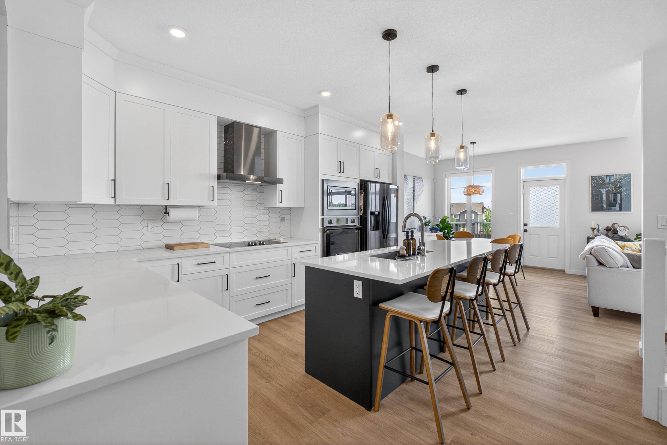 Modern kitchen featuring white cabinetry, a dark-toned island with seating, a stainless steel range hood, integrated appliances, and light wood-finish flooring - 17023 45 Street, Edmonton, AB