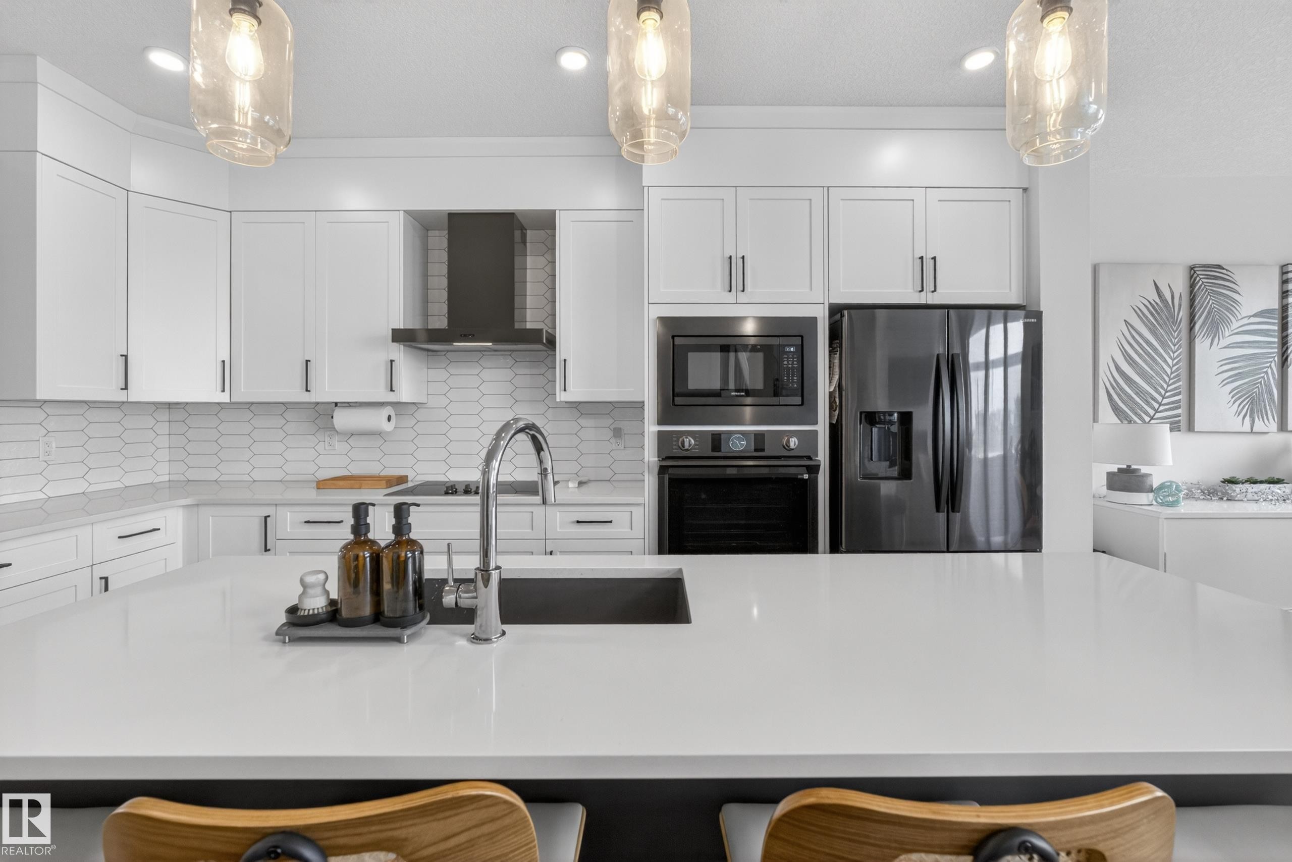 Modern kitchen featuring white cabinetry, a large center island with an undermount sink, stainless steel appliances, a range hood, and a white picket tile backsplash - 17023 45 Street, Edmonton, AB