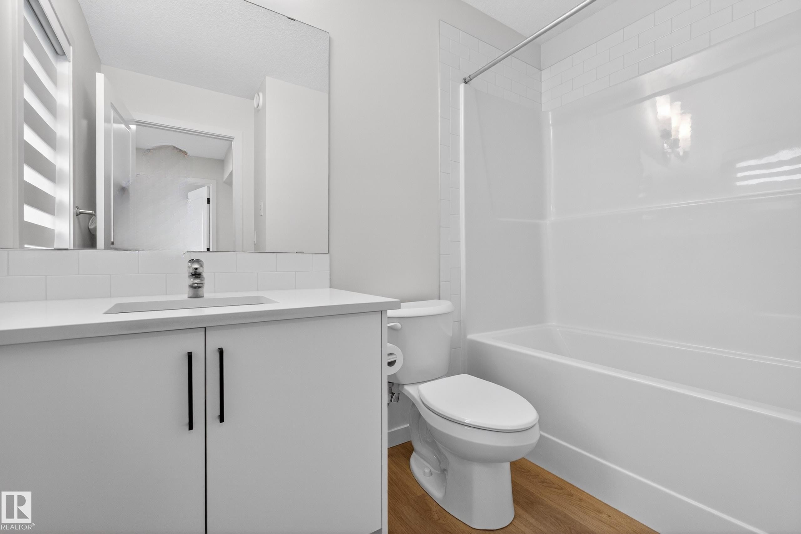 Bathroom featuring a white vanity with countertop sink, large wall-mounted mirror, modern black hardware, wood-finish flooring, and a white tiled bathtub with shower rod - 17023 45 Street, Edmonton, AB