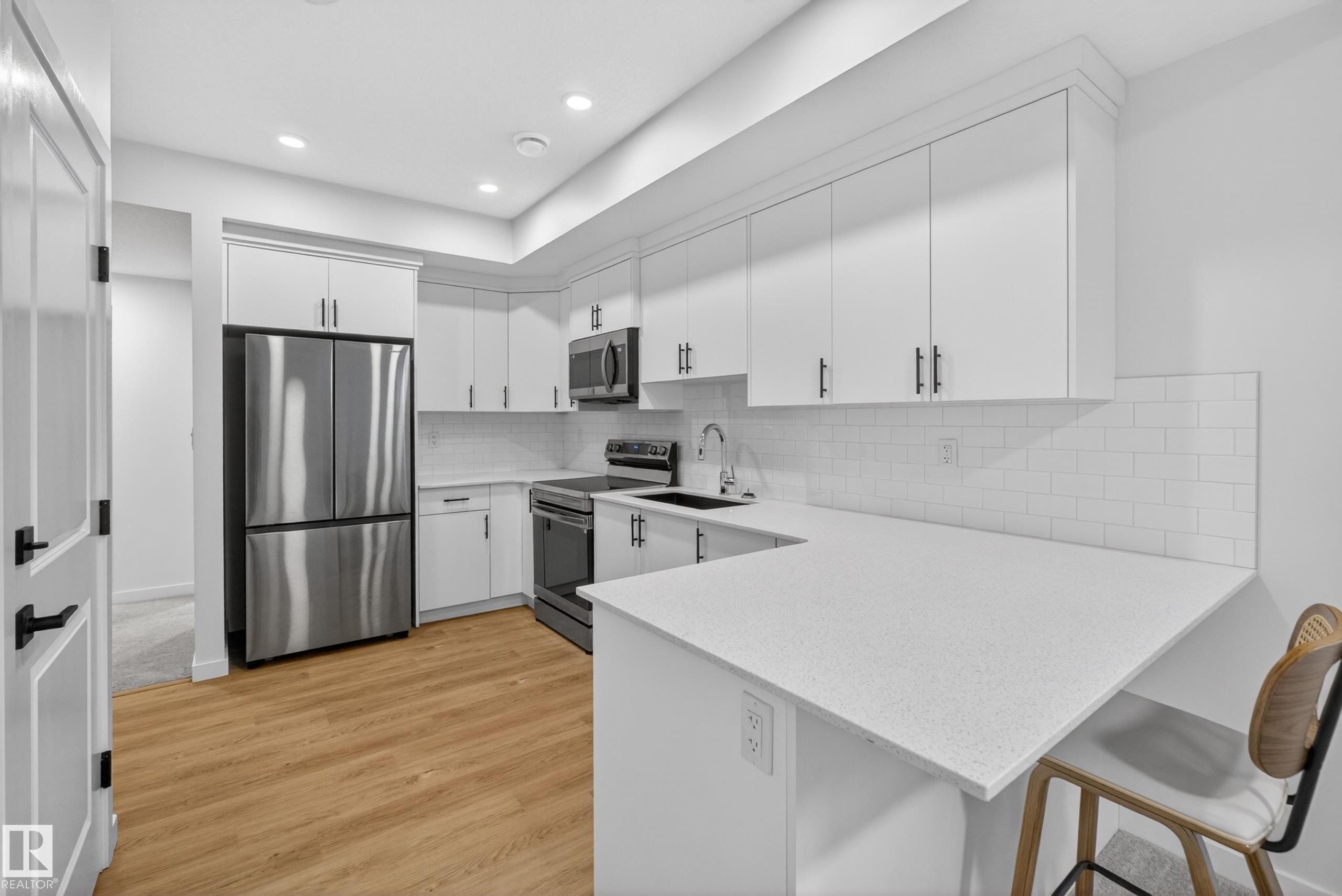 Modern kitchen featuring white cabinetry, stainless steel appliances, a white subway tile backsplash, light-toned wood-finish flooring, and a stone-finish countertop with an integrated breakfast bar - 17023 45 Street, Edmonton, AB