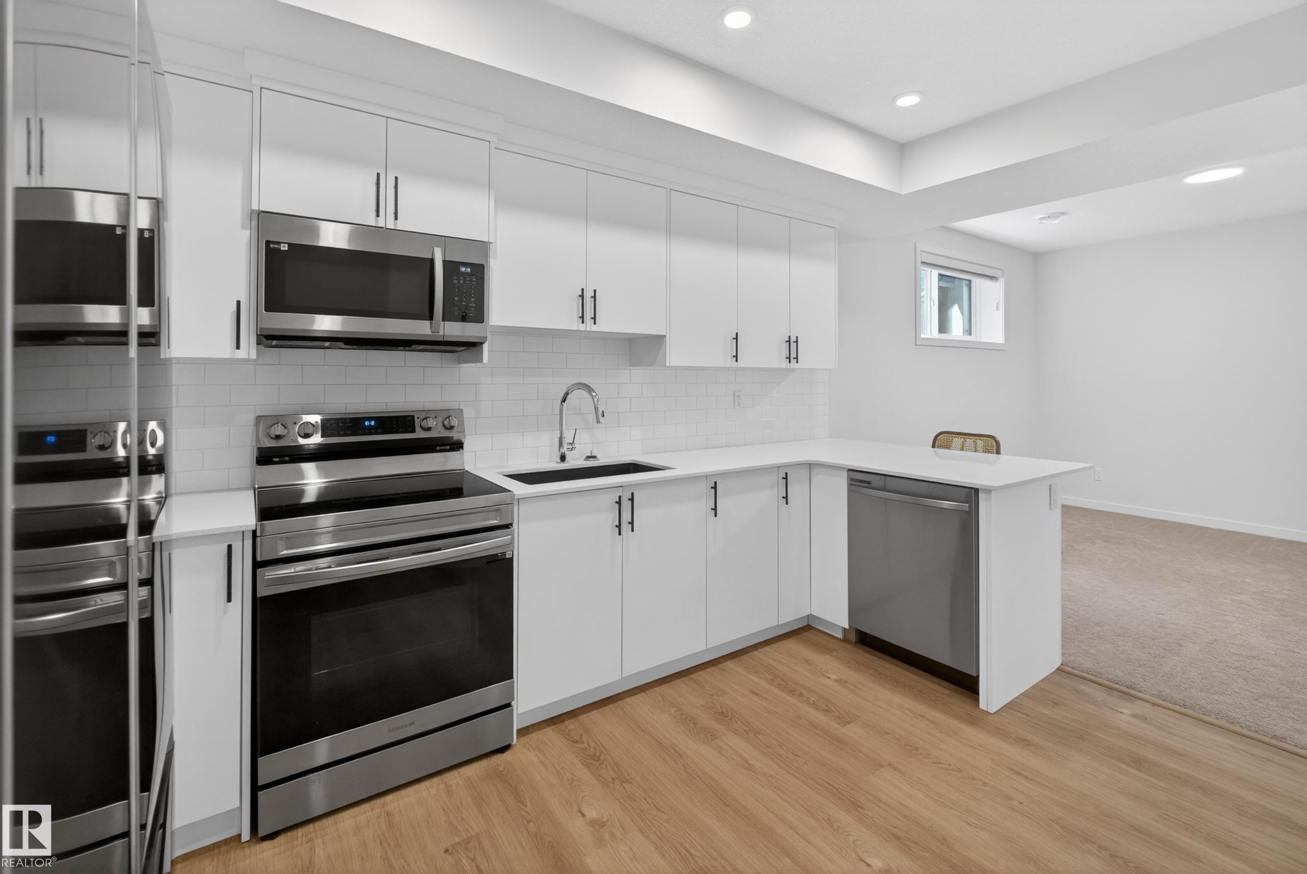 Modern kitchen featuring white cabinetry, stainless steel appliances, a subway tile backsplash, a dark basin sink, and wood-finish flooring - 17023 45 Street, Edmonton, AB