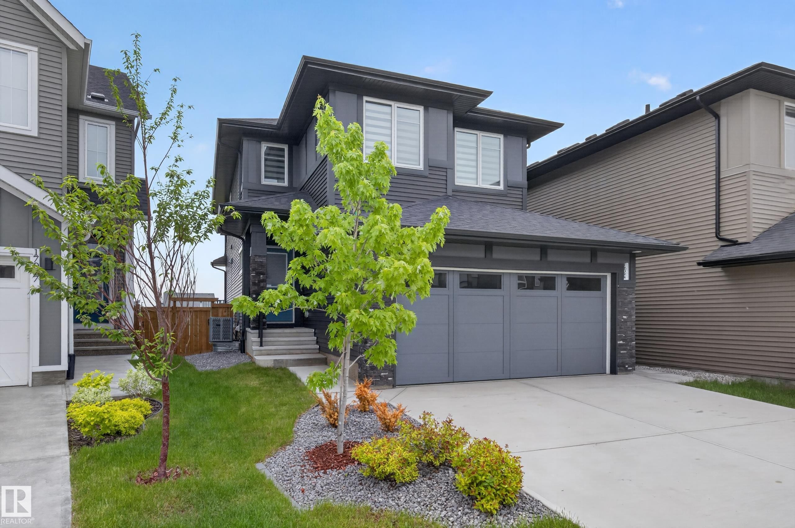 Contemporary two-story residence with a dark gray exterior, featuring a two-car attached garage, concrete driveway, and front landscaping with mature trees and shrubs - 17023 45 Street, Edmonton, AB