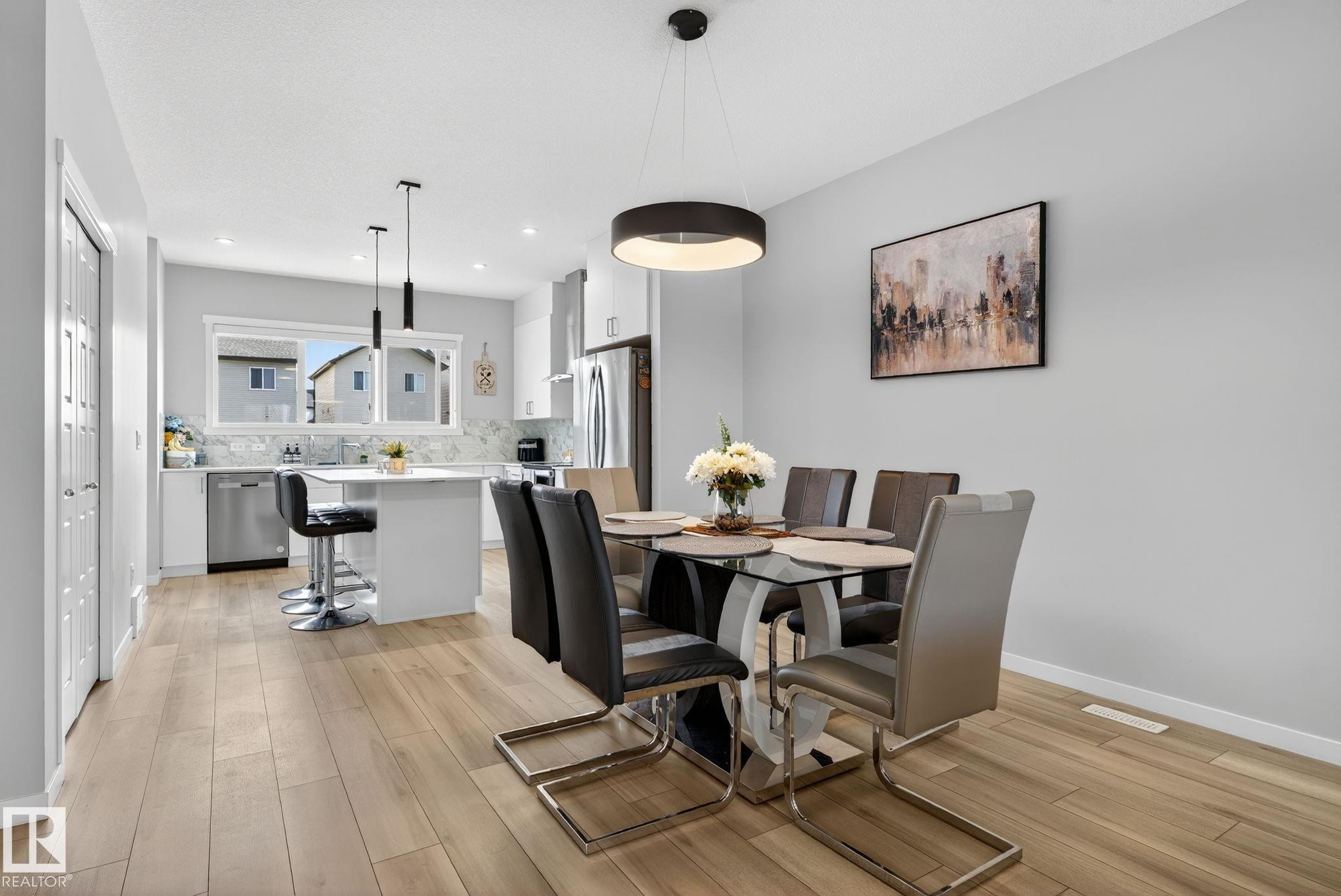 Open-concept living space featuring wood-finish flooring, a contemporary dining area with a circular pendant light fixture, and a kitchen with white cabinetry, stainless steel appliances, and a central island with pendant lighting - 1213 16A Street, Edmonton, AB