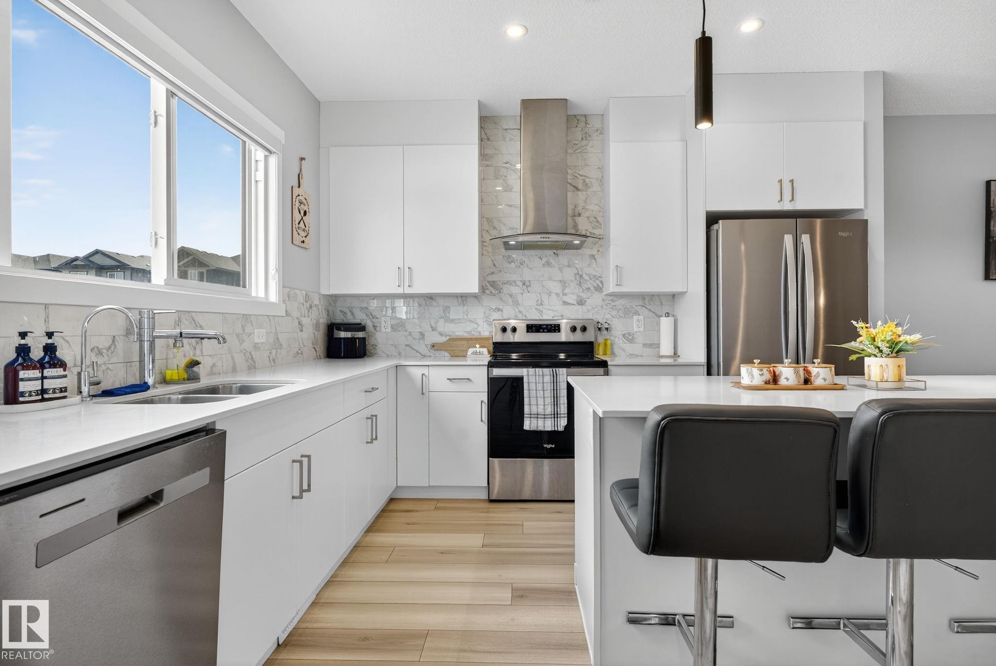 Contemporary kitchen featuring white cabinetry, stainless steel appliances, a white quartz countertop island with seating, and a marble-patterned backsplash - 1213 16A Street, Edmonton, AB