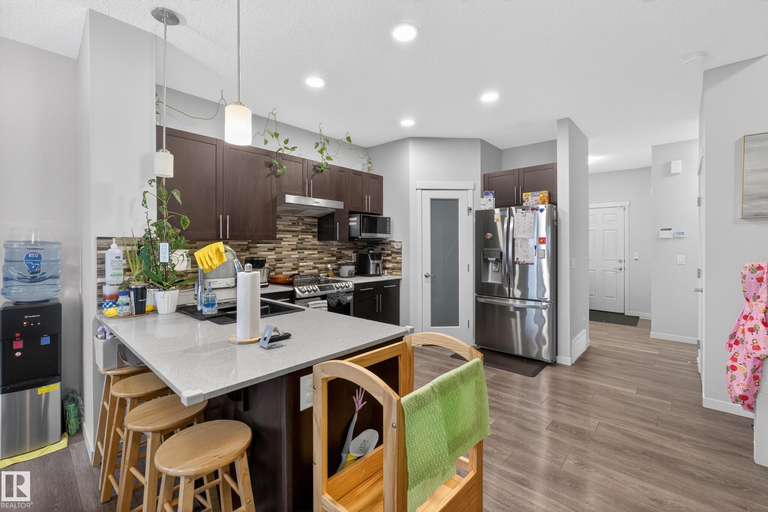 Open-concept kitchen featuring dark wood-finish cabinetry, a light-toned countertop island with pendant lighting, a mosaic tile backsplash, stainless steel appliances, and wood-finish flooring - 1404 25 Avenue, Edmonton, AB
