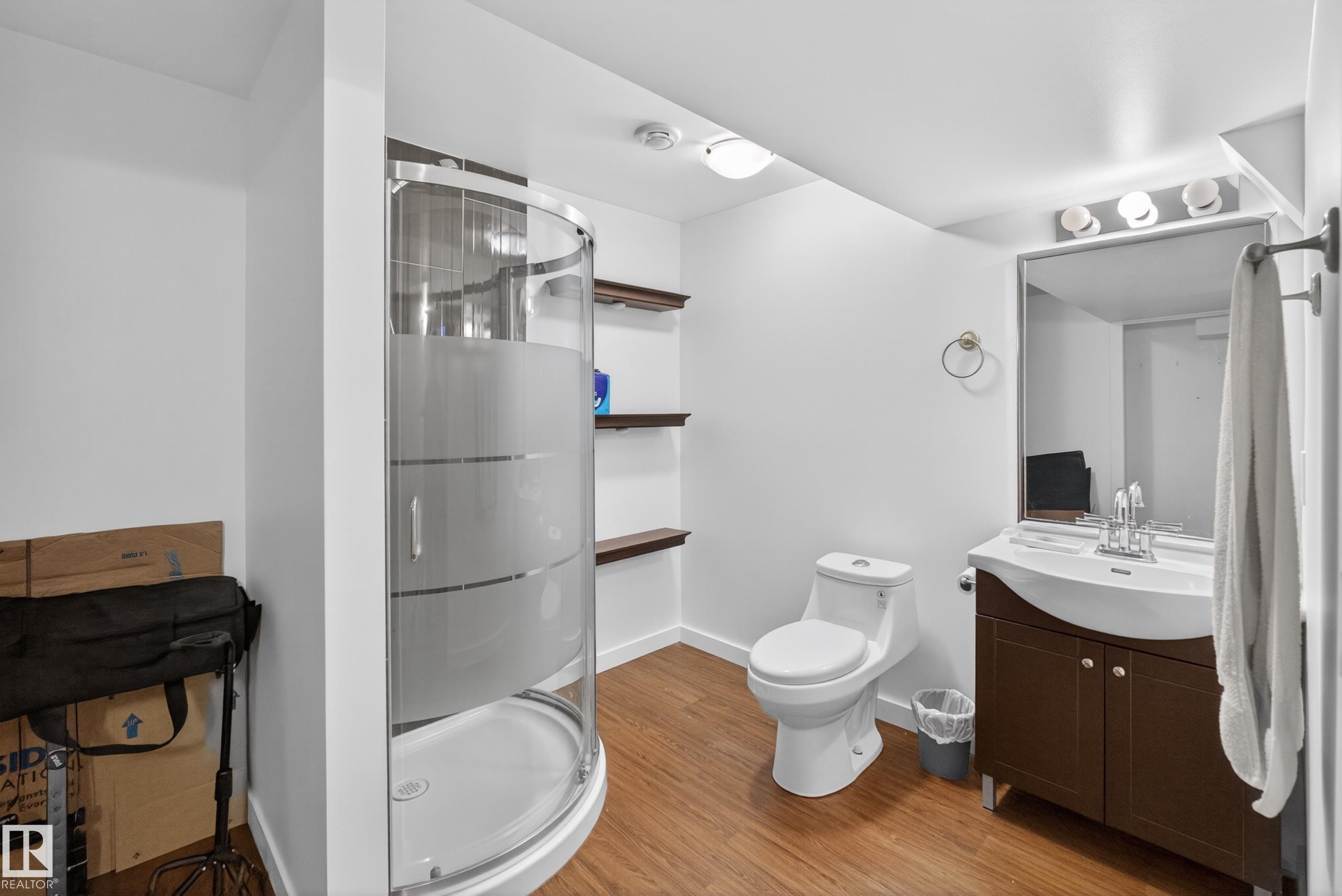 Full bathroom featuring a curved glass shower enclosure, wood-finish flooring, a vanity with an integrated sink, a rectangular mirror with overhead lighting, and wall-mounted shelving - 1404 25 Avenue, Edmonton, AB