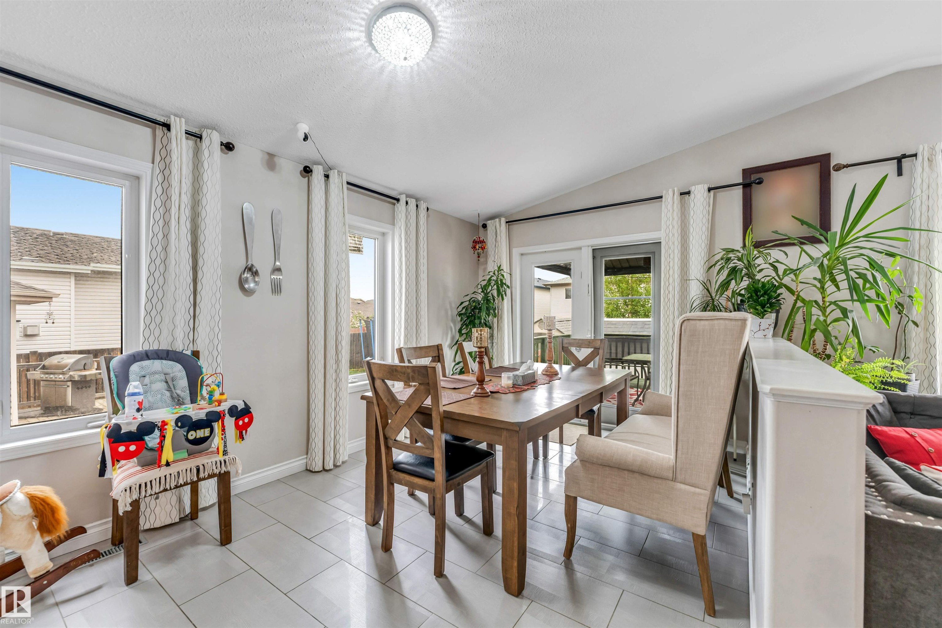 Dining area featuring light gray tile flooring, multiple windows with white curtains, a white ceiling fan light fixture, and a glass-paneled door leading to an outdoor space - 3519 22 Street Nw, Edmonton, AB