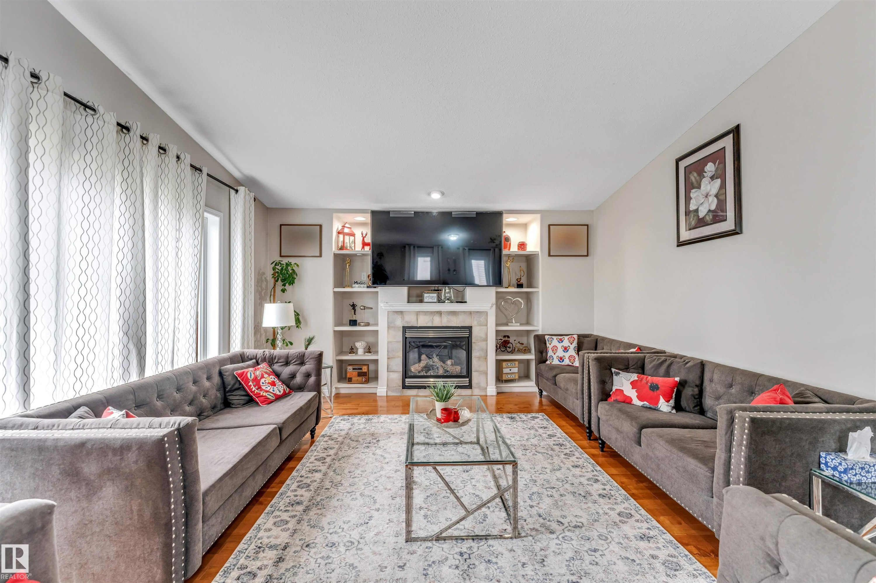 Living room featuring hardwood-finish flooring, a central fireplace with stone surround, integrated shelving with accent lighting, and large windows - 3519 22 Street Nw, Edmonton, AB