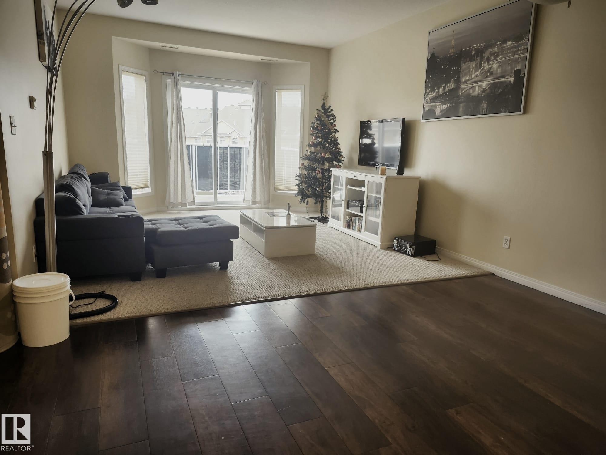 Spacious living area featuring bay windows, a glass-paneled balcony door, wood-finish flooring, and light-toned wall paint - 425 4304 139 Avenue, Edmonton, AB