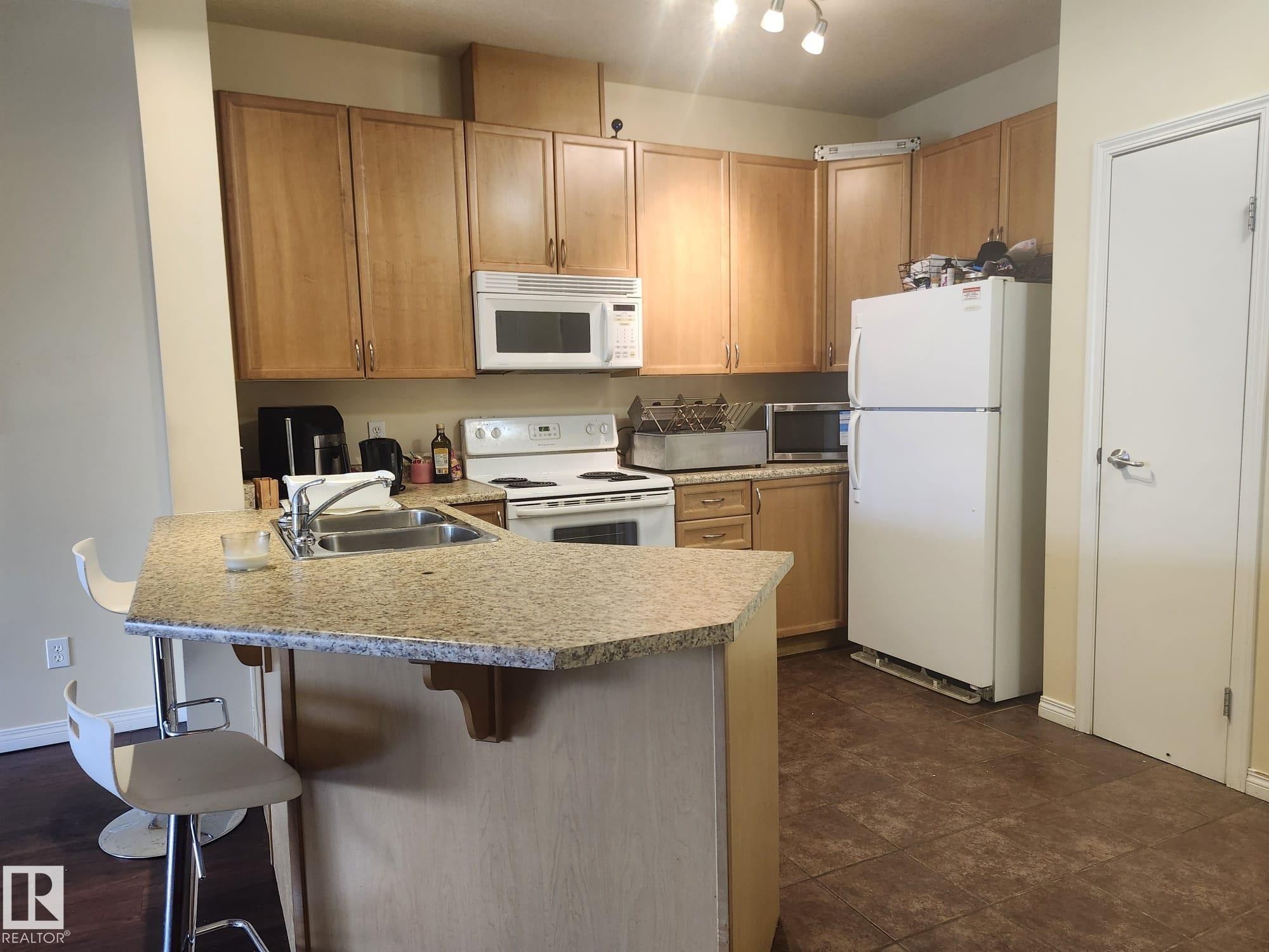 Kitchen featuring a granite-look countertop island with an undermount sink, overhead track lighting, wood cabinetry, and a tile-look flooring - 425 4304 139 Avenue, Edmonton, AB