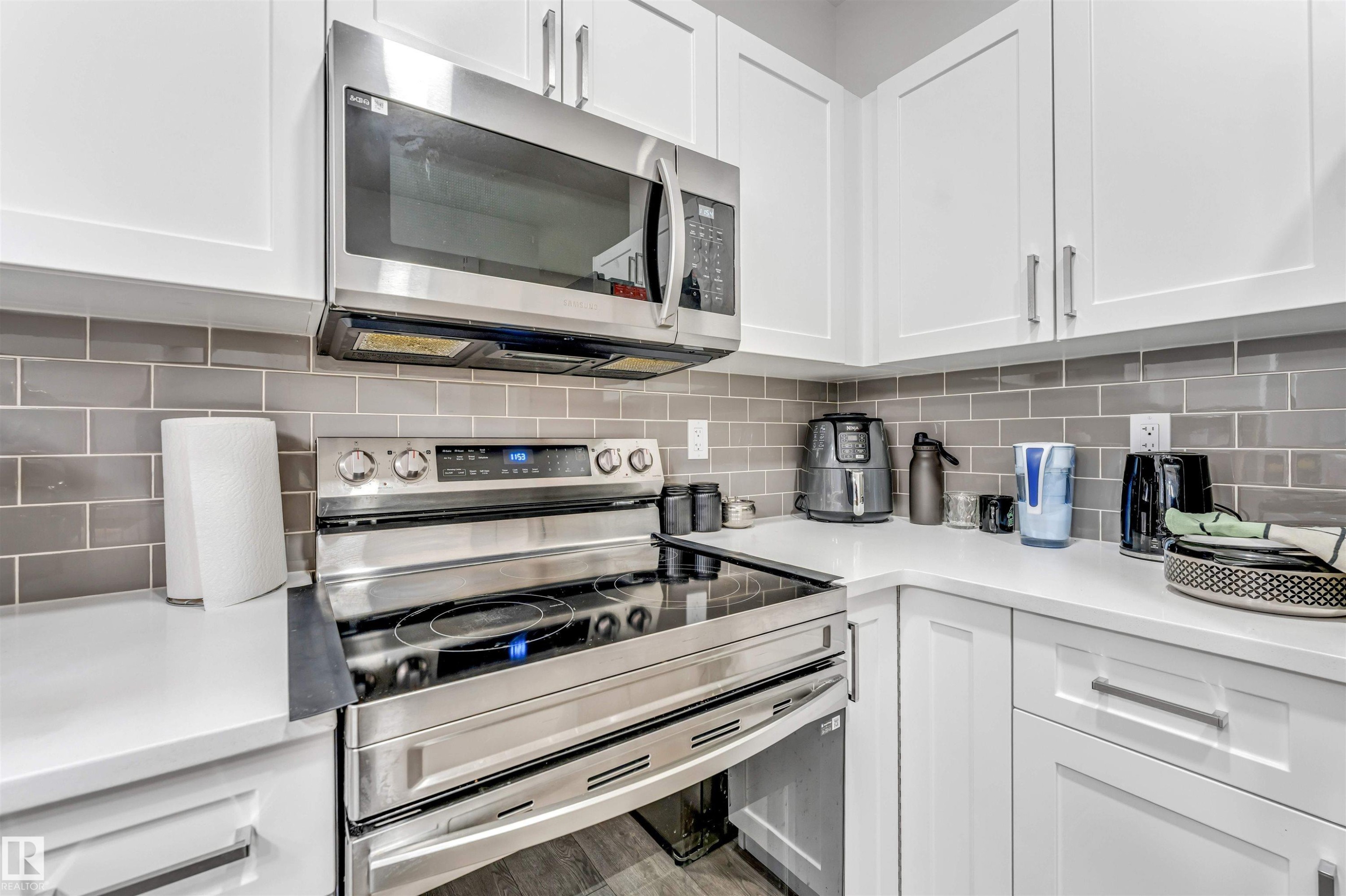 Corner kitchen featuring white upper and lower cabinetry, stainless steel range with electric cooktop, stainless steel over-the-range microwave, gray subway tile backsplash, and light-toned countertops - 45 2710 66 Street, Edmonton, AB