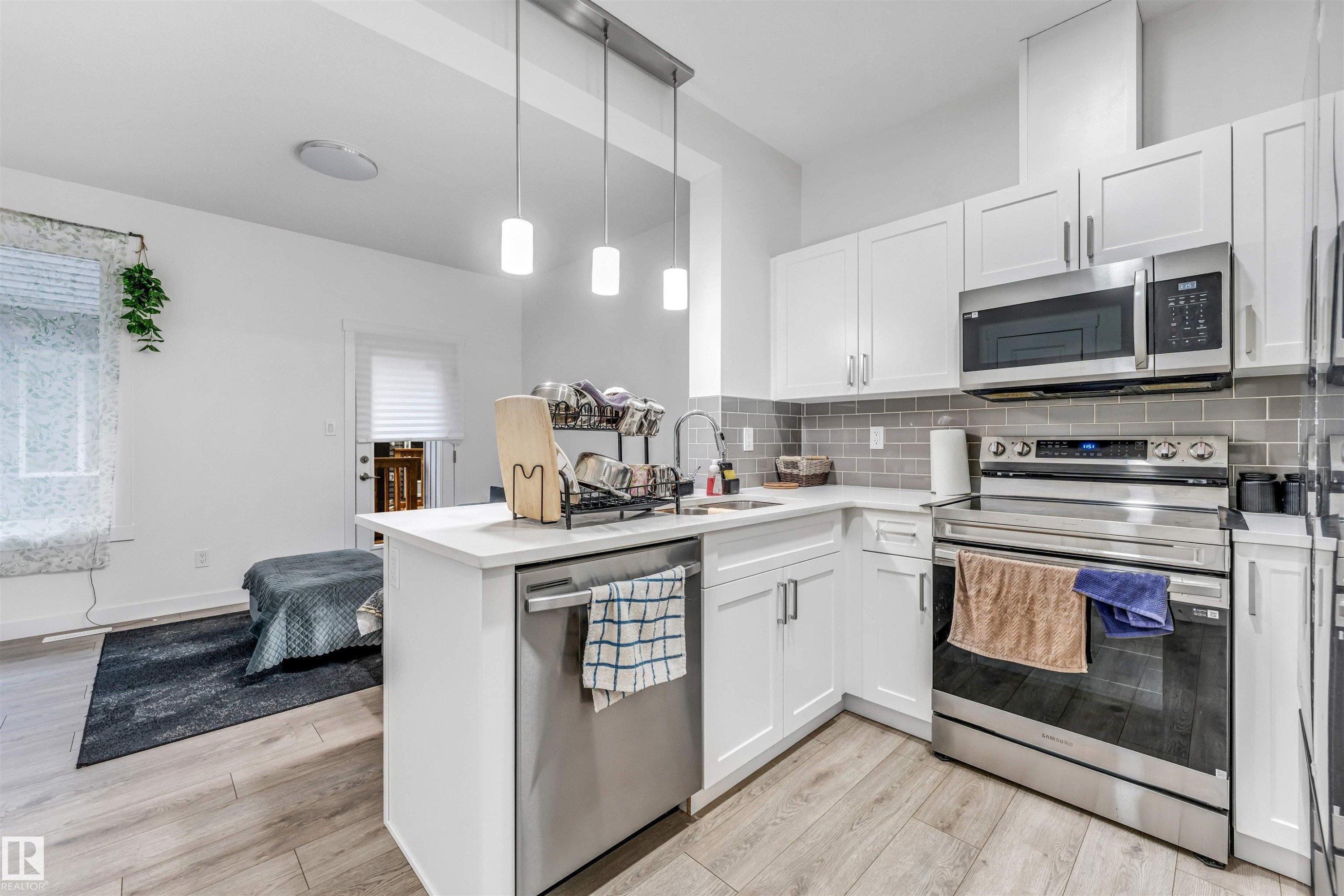 Modern kitchen featuring white shaker cabinetry, stainless steel appliances, light wood-finish flooring, a white countertop island with a built-in sink, and a gray subway tile backsplash - 45 2710 66 Street, Edmonton, AB