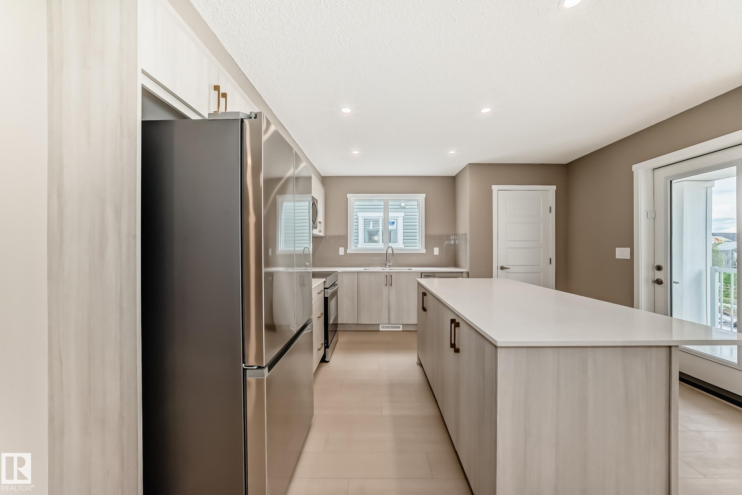 Modern kitchen featuring a large central island with a light-colored countertop, stainless steel appliances, light wood-finish cabinetry, recessed lighting, and a sliding glass door leading to a balcony - 1210 Stoneside Road, Sherwood Park, AB - Indoor Photo Showing Kitchen With Upgraded Kitchen