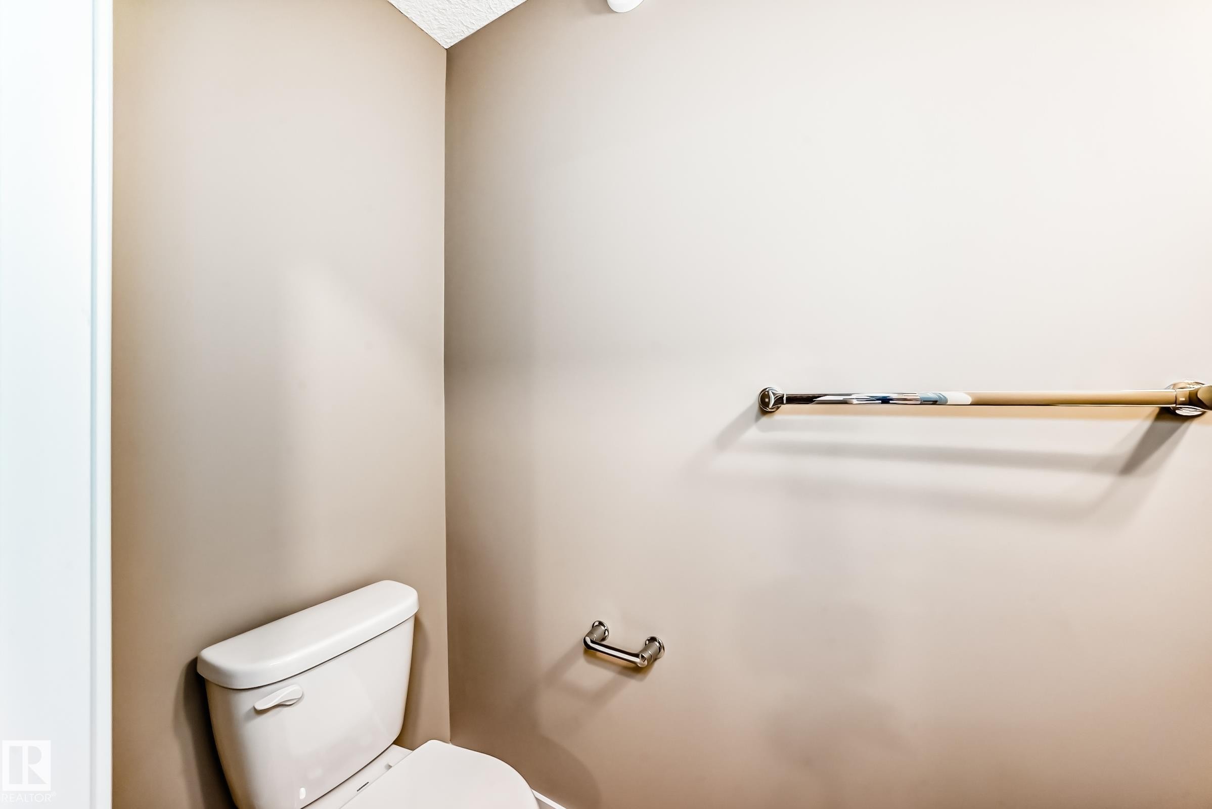 White toilet with a beige painted wall, textured ceiling, chrome toilet paper holder, and a wooden grab bar with chrome mounts - 1210 Stoneside Road, Sherwood Park, AB - Indoor Photo Showing Bathroom