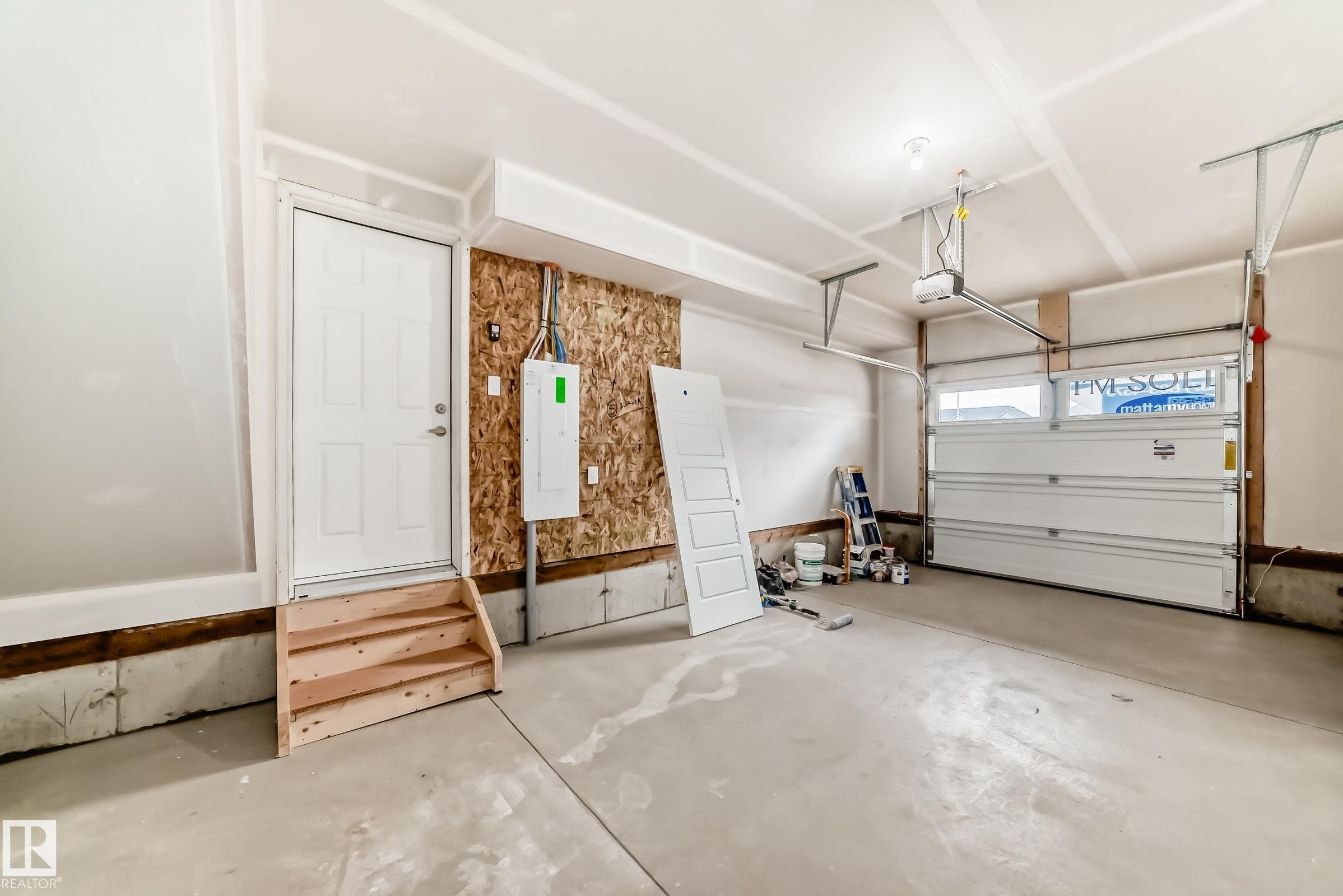 Spacious garage featuring a concrete floor, an overhead garage door with an automatic opener, exposed OSB wall sheathing, and a service entry door with wooden steps - 1210 Stoneside Road, Sherwood Park, AB - Indoor Photo Showing Garage